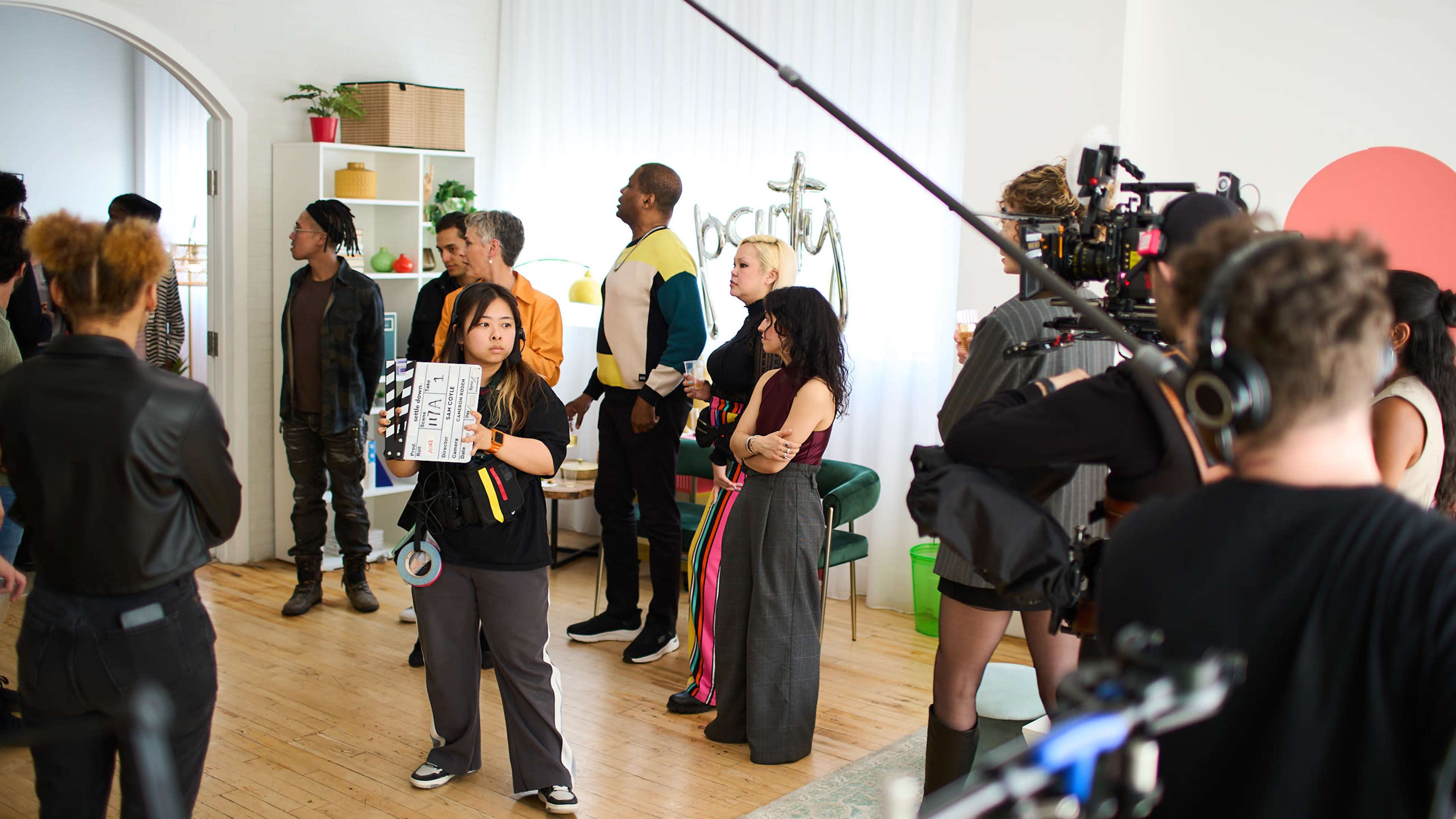 A diverse group of people is gathered on a film set, with one person holding a clapperboard and a camera crew preparing for a shot.