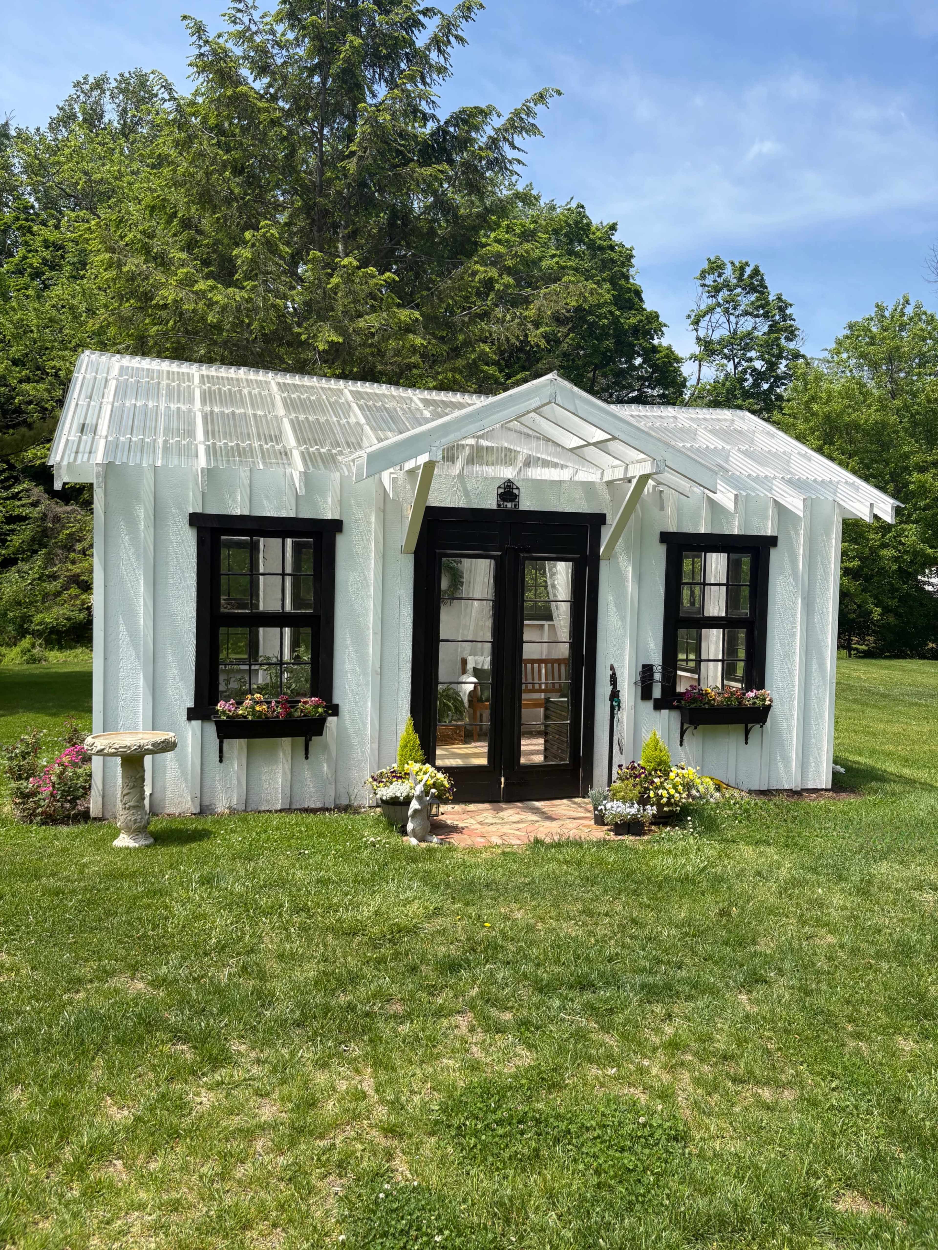 A small white cottage with a transparent roof sits in a grassy yard, surrounded by flower beds and decorative elements.
