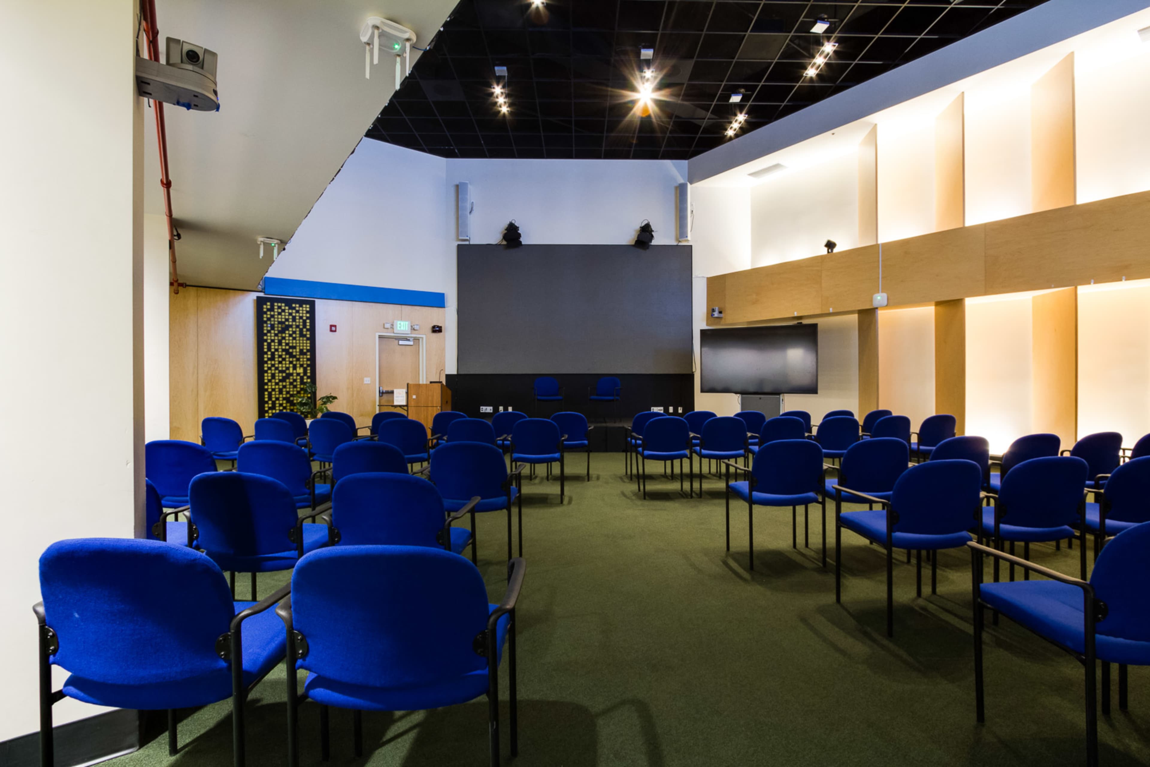 A conference room with blue chairs arranged in rows facing a podium and a screen on a stage.