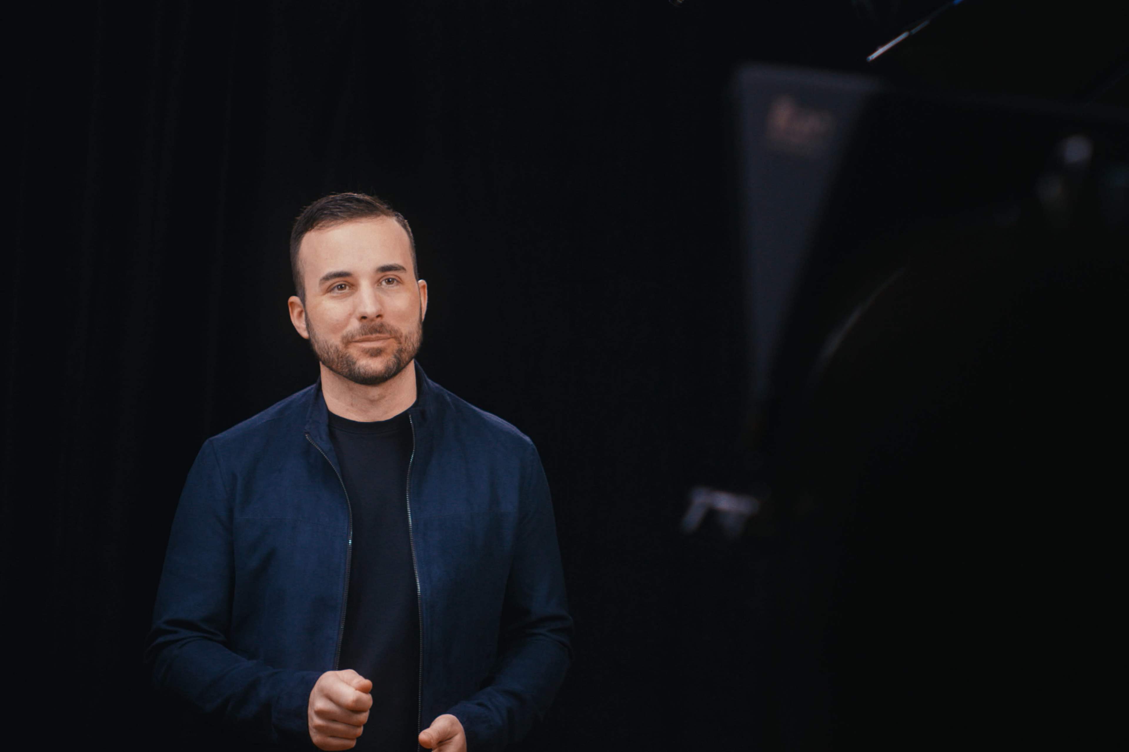 A man in a blue jacket stands in front of a dark backdrop, speaking energetically into a camera.