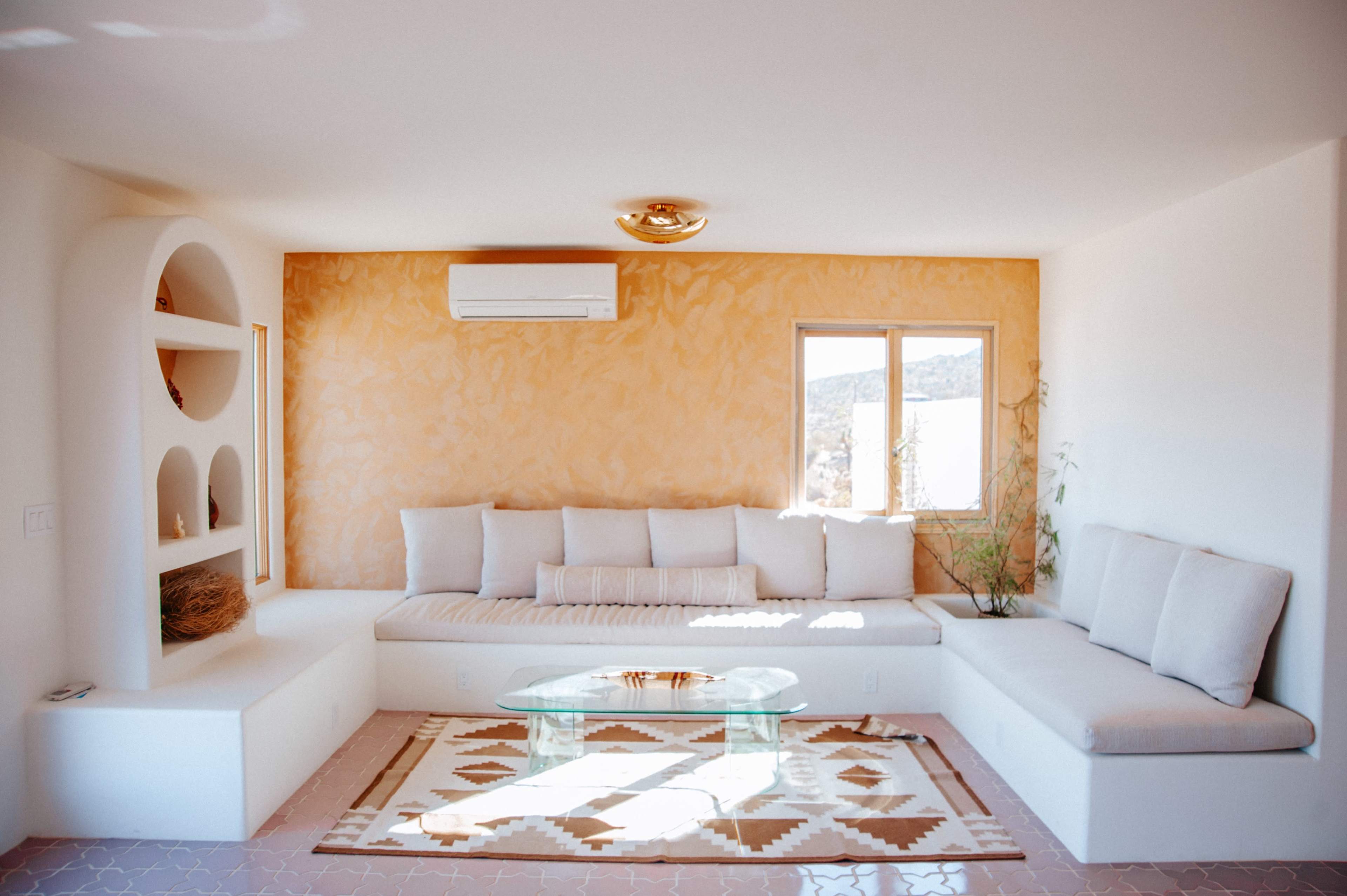 A modern living room with a beige sectional sofa, a glass coffee table, and decorative wall shelves against a light orange wall.