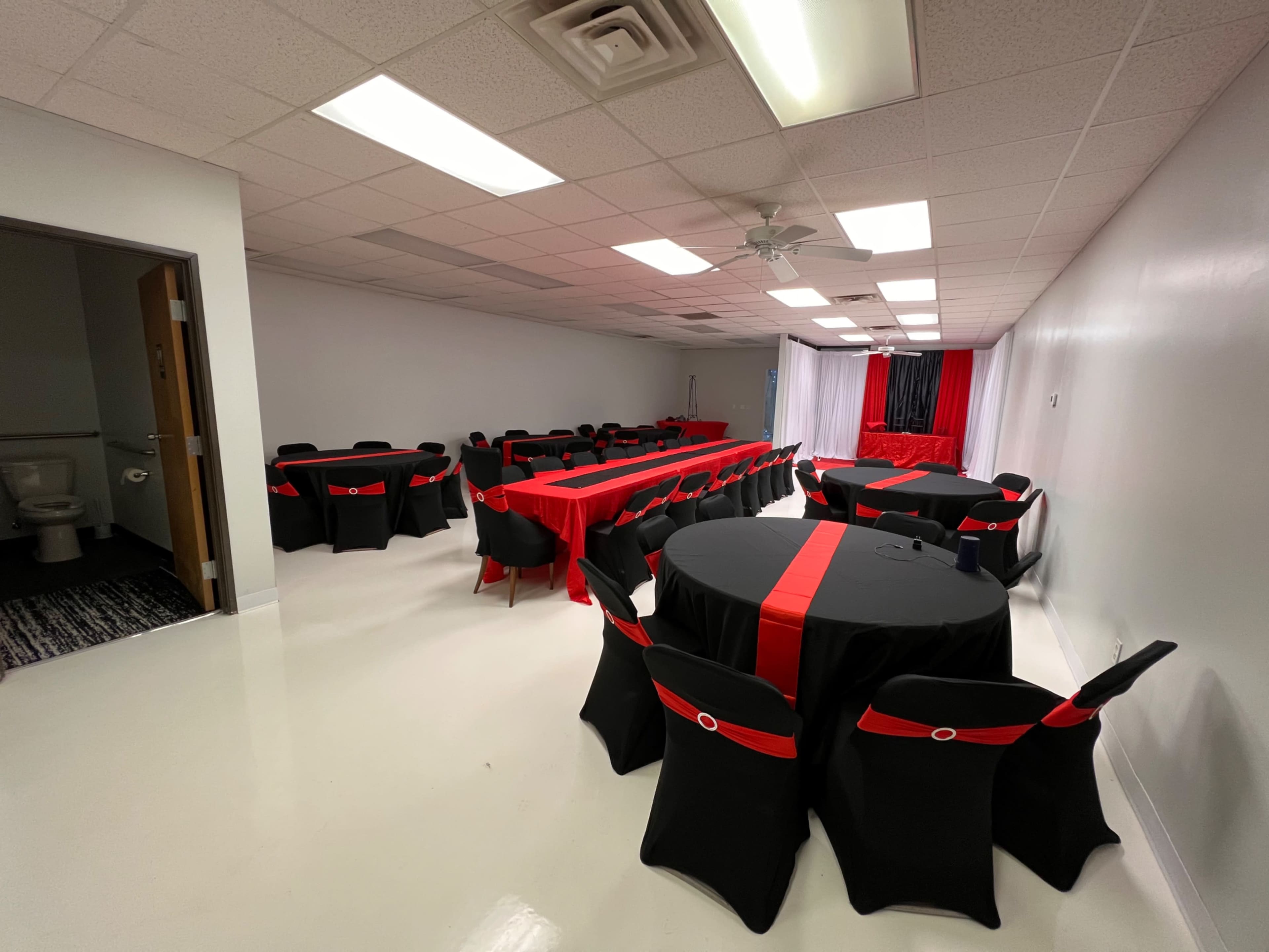 A banquet hall set up with round tables covered in black tablecloths adorned with red ribbons, illuminated by overhead fluorescent lights.