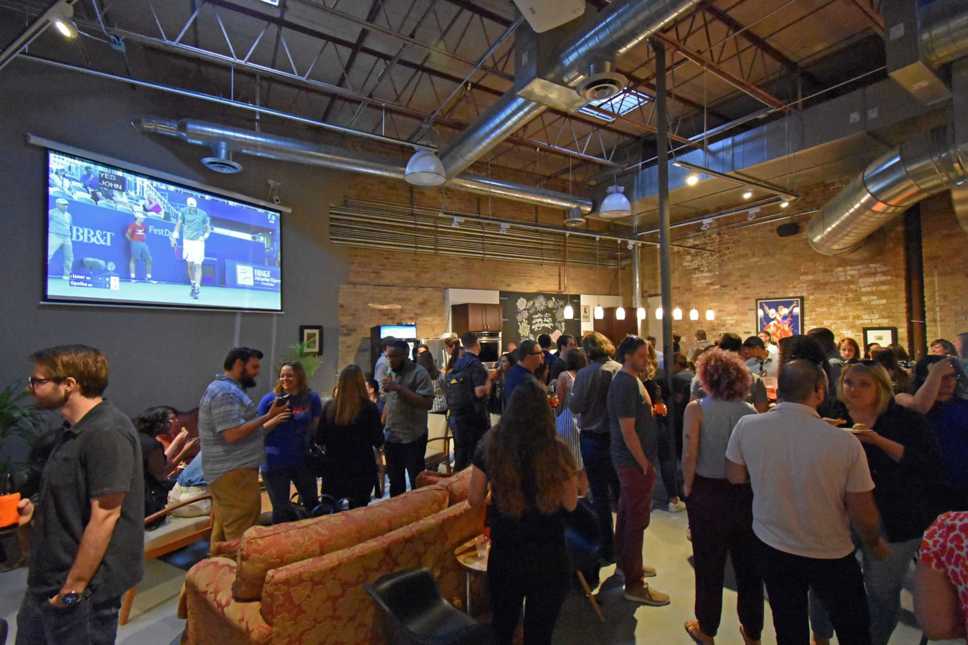 A crowd gathers in a spacious lounge area with exposed brick walls, watching a tennis match broadcast on a large screen.