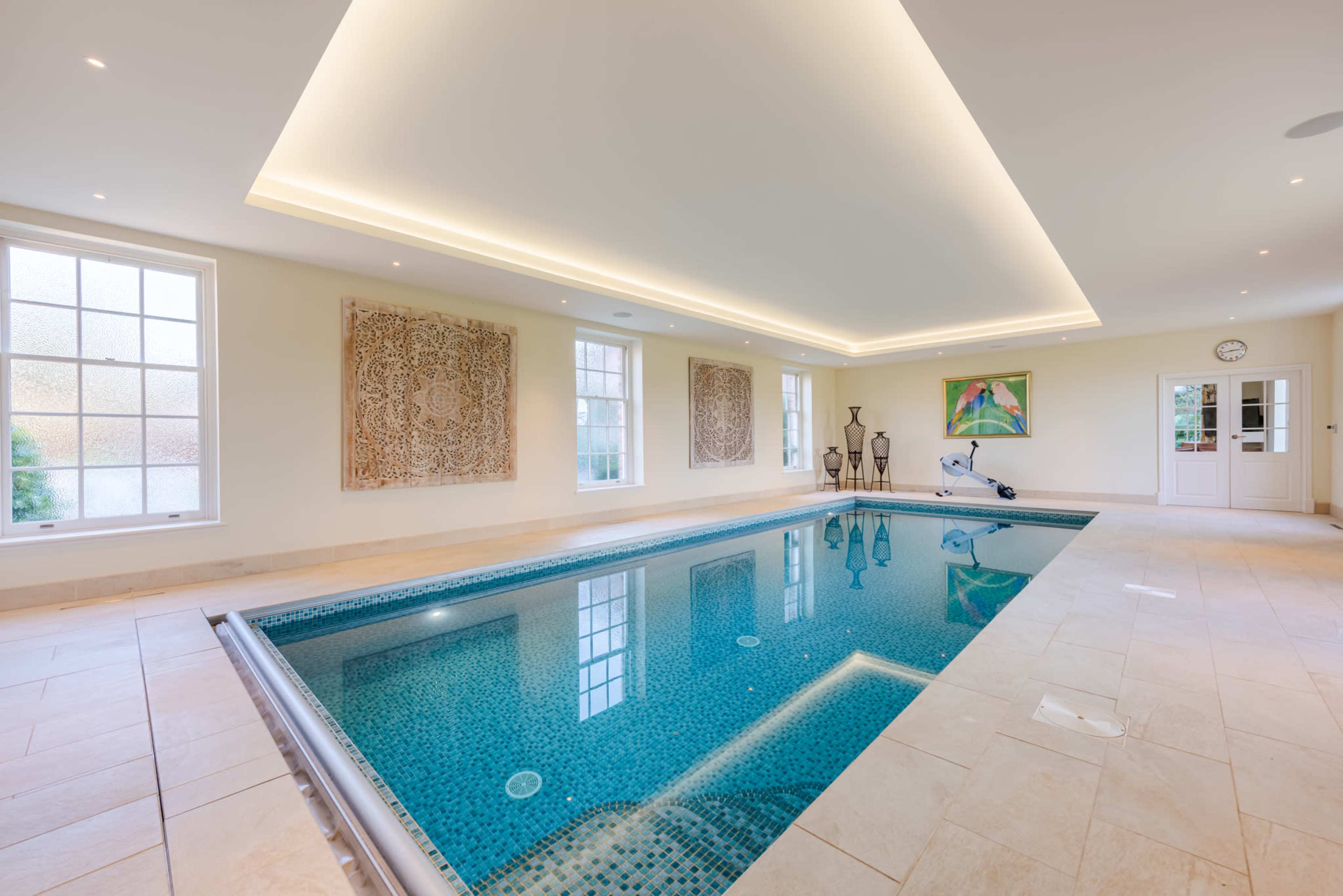 The image shows an indoor pool area with tiled flooring, elegant lighting, and decorative wall art surrounding the pool.