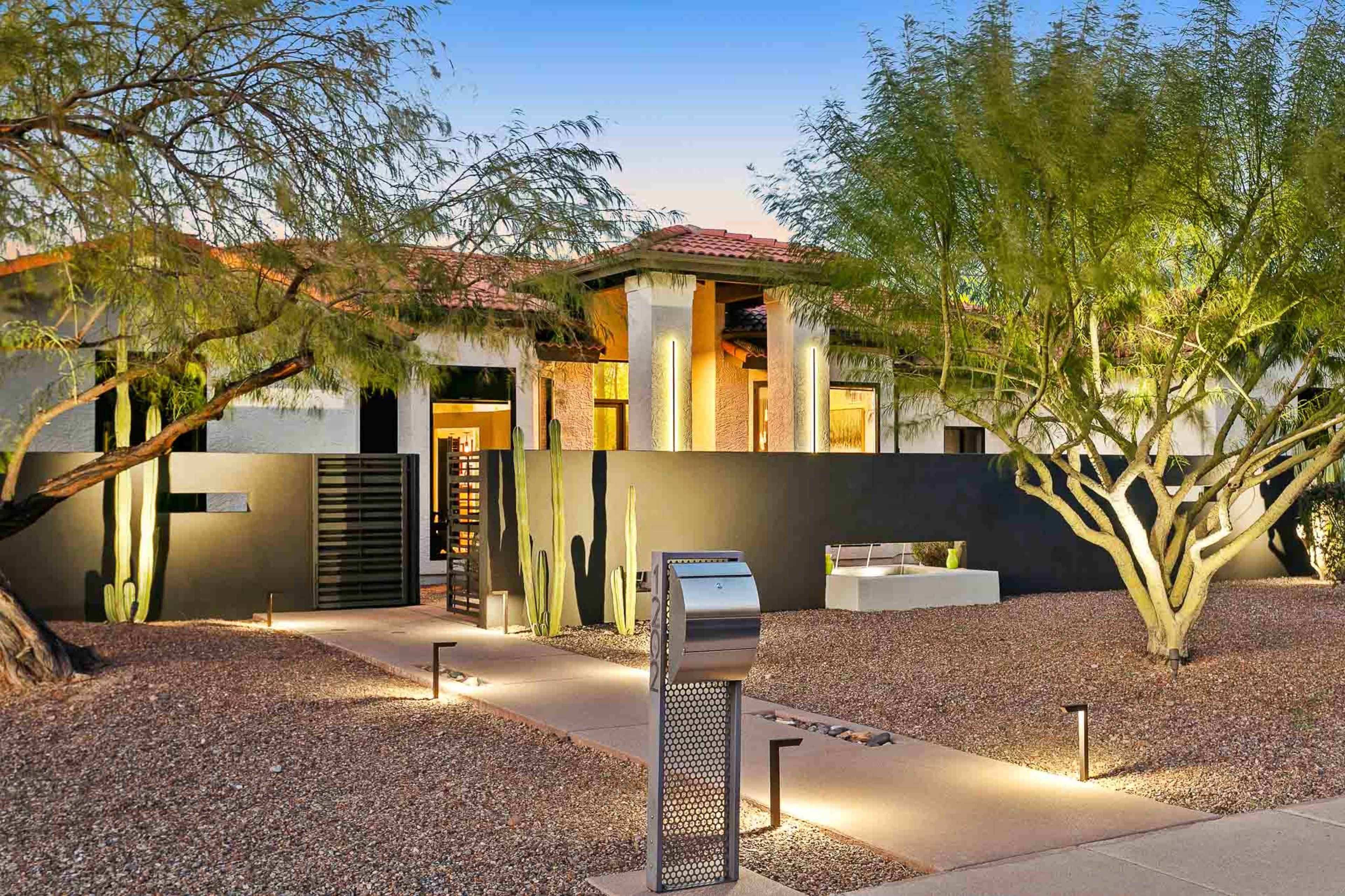 The image shows a modern single-story home surrounded by desert landscaping, featuring a pathway lined with lights and a mailbox near the entrance.