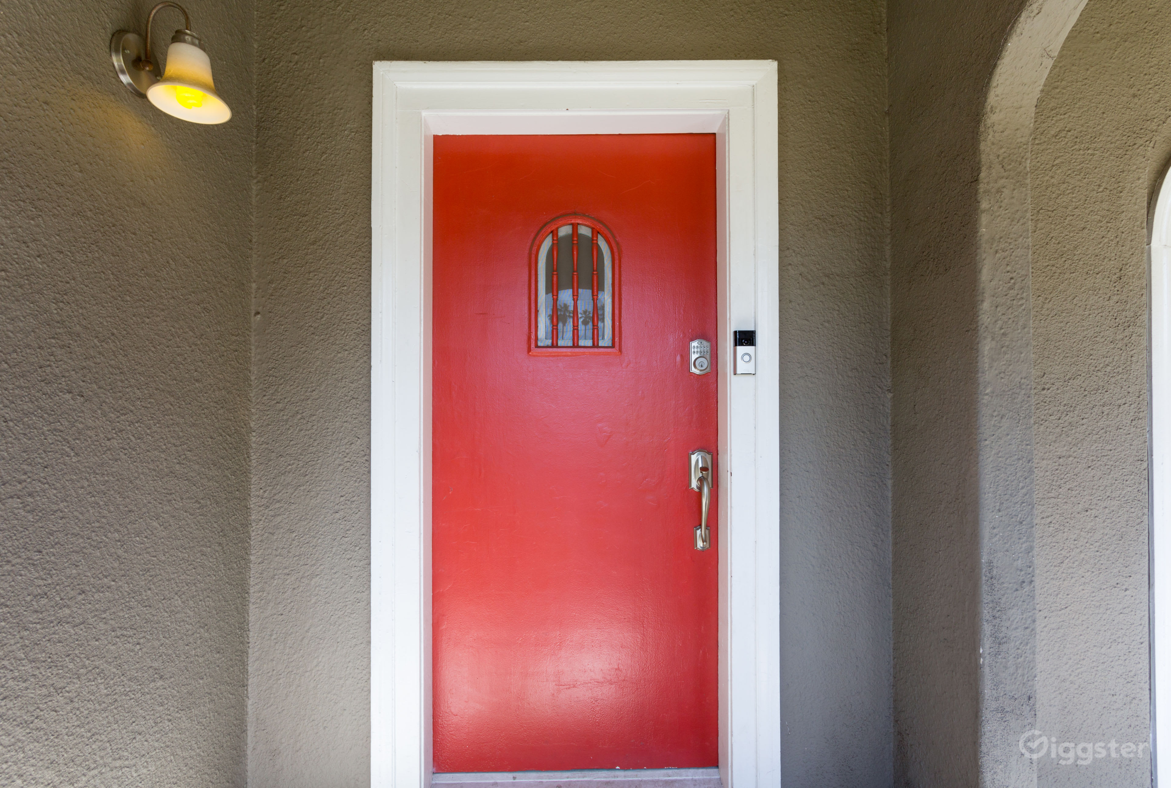 The image shows a red front door with a decorative window, framed by a light gray wall and a small overhead light.