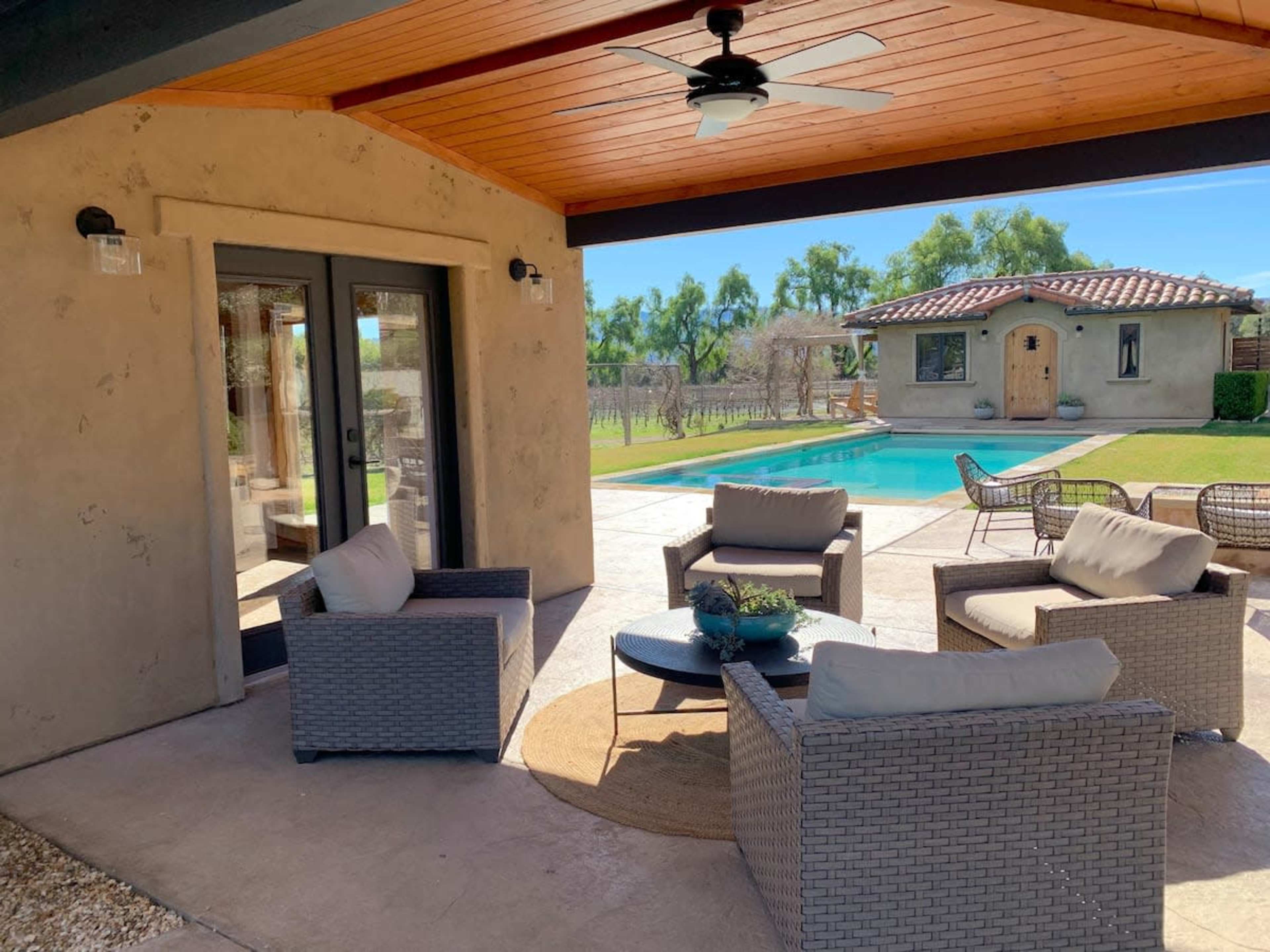 A shaded outdoor lounge area features four chairs around a circular table, adjacent to a swimming pool and a rustic building in a landscaped yard.