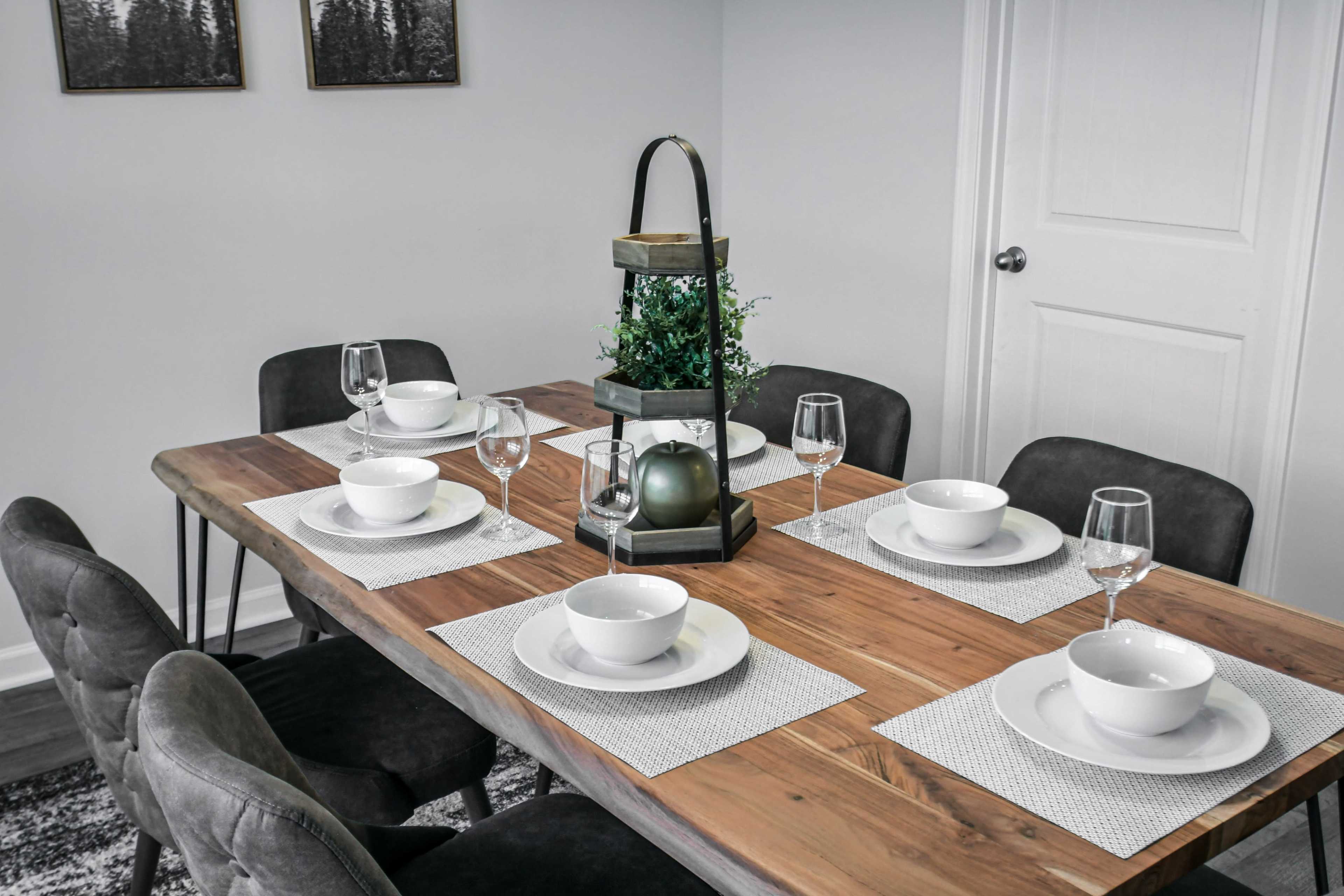 A wooden dining table is set with white plates and glassware, surrounded by six gray chairs, and featuring a lantern centerpiece with greenery.