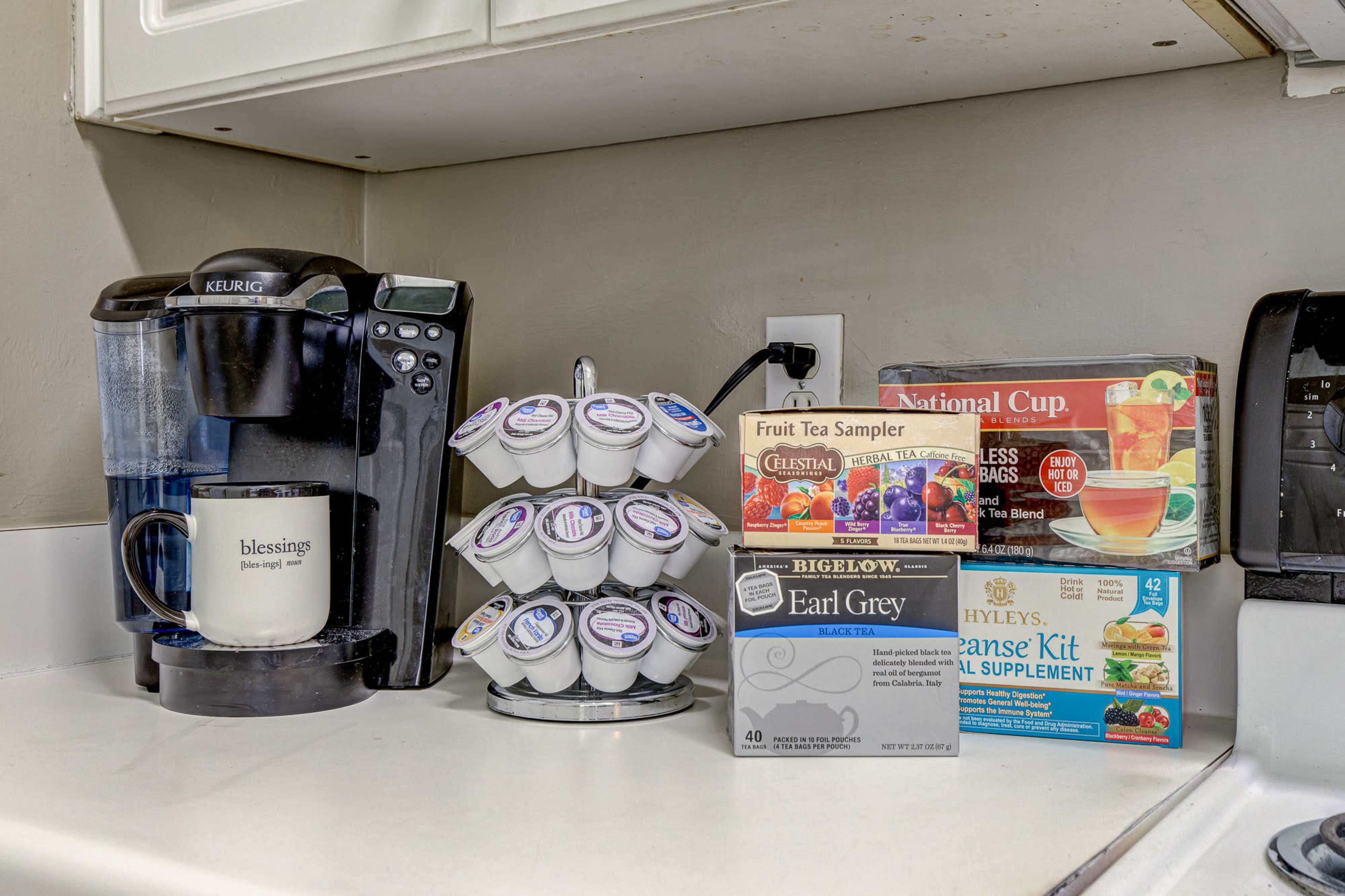 A coffee maker sits beside a rack of assorted tea pods, with boxes of tea and a mug on a kitchen counter.