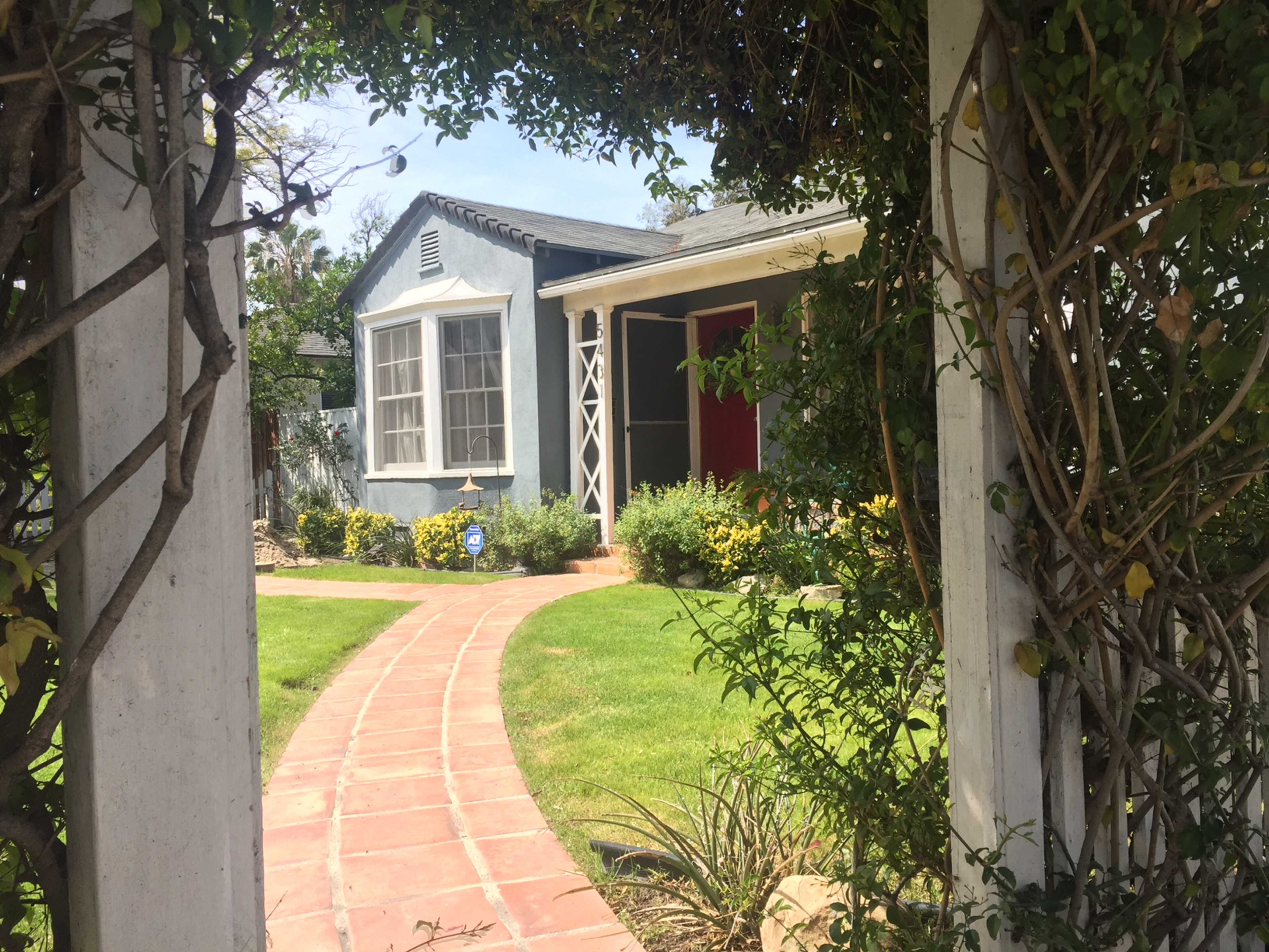 A pathway leads to a small, blue house with a red door, framed by a trellised archway covered in greenery.