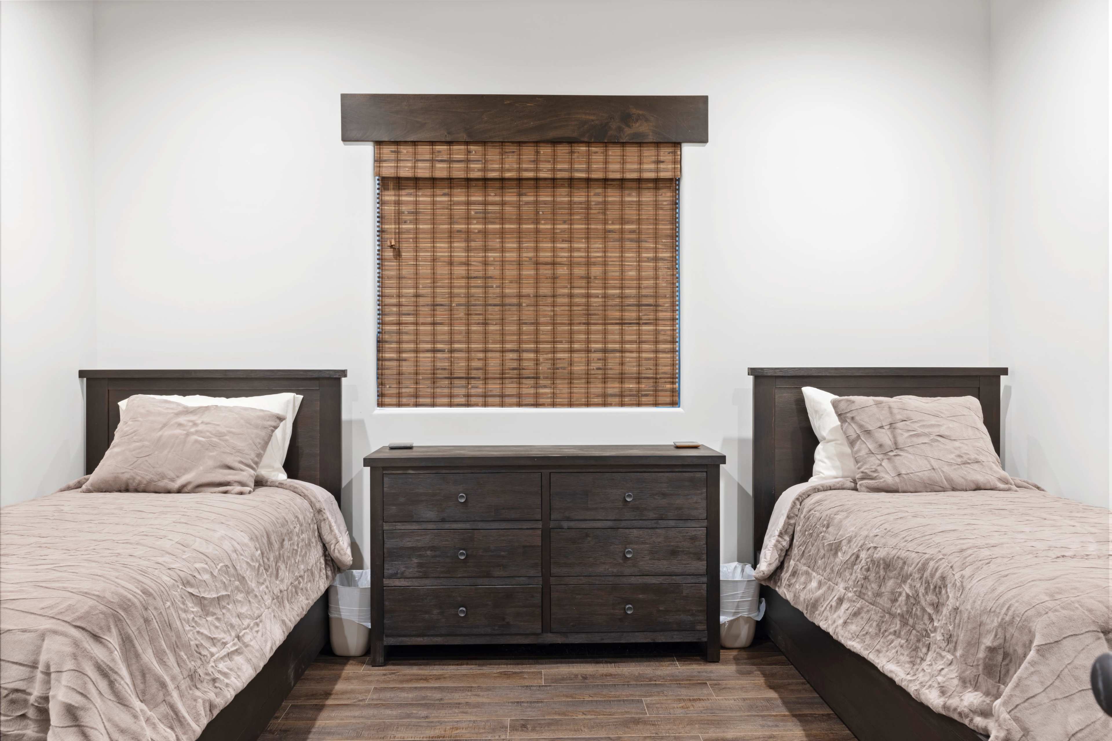 The image shows a minimalist bedroom with two twin beds, a wooden dresser in the center, and a window covered by a bamboo blind.