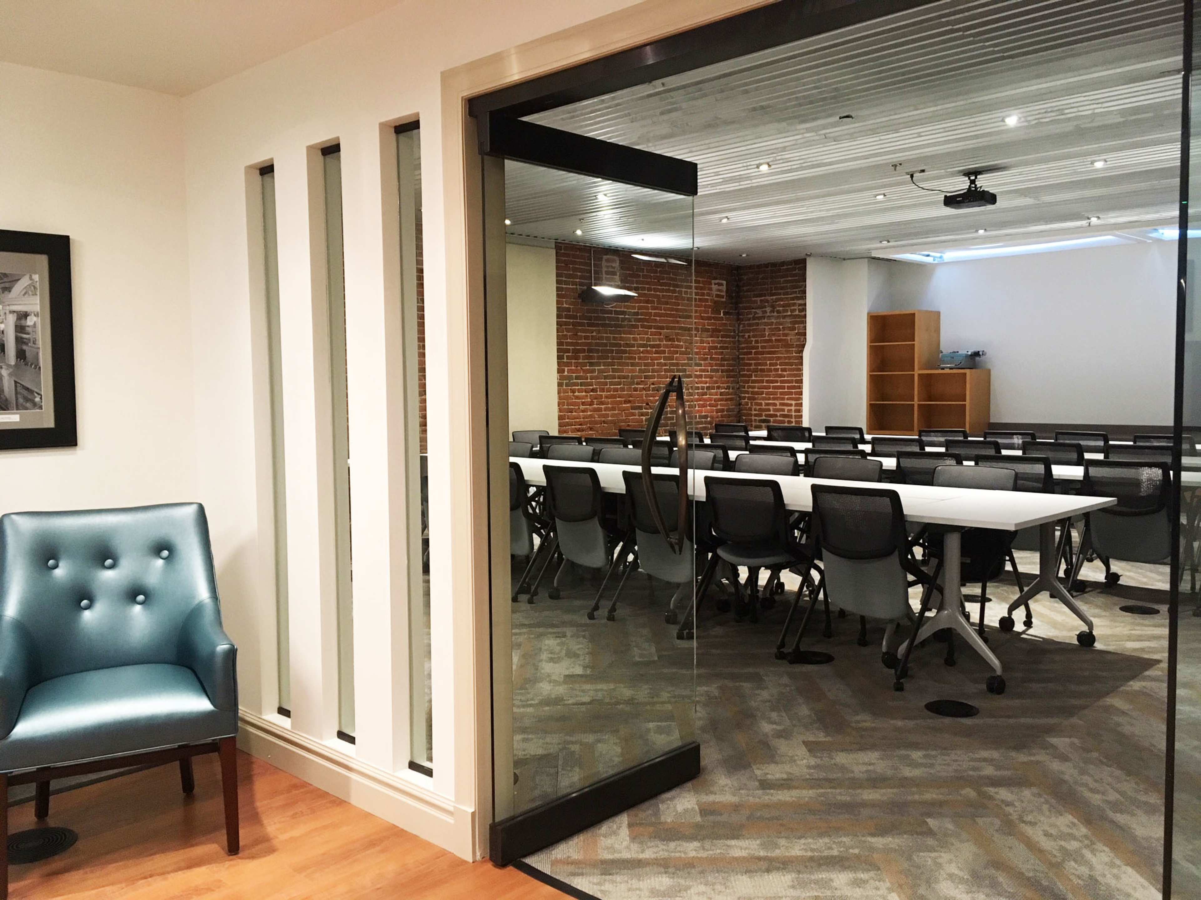 A meeting room features a large, empty table surrounded by modern chairs, with a view through a glass door from a hallway.