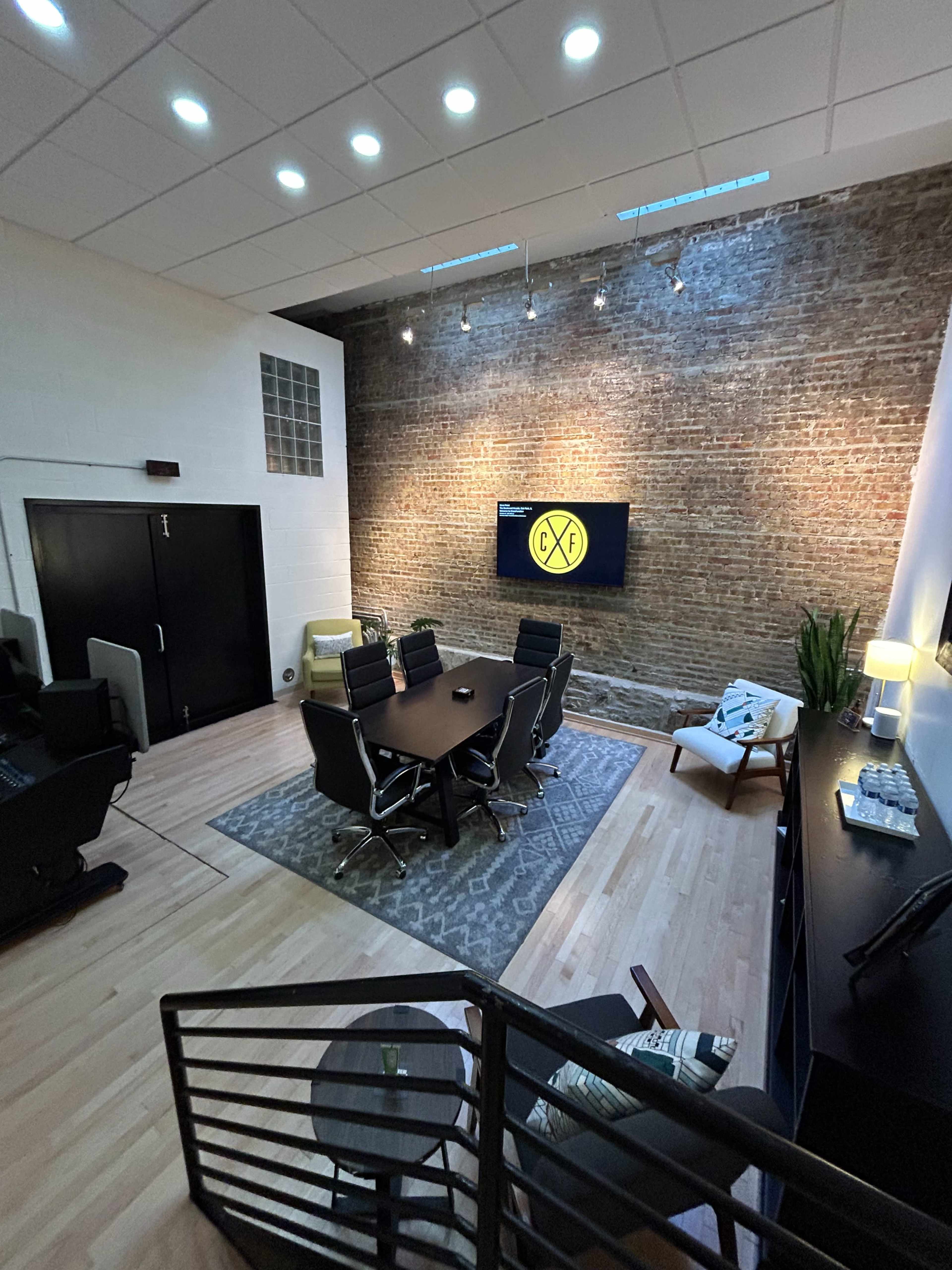A modern office space featuring a conference table, ergonomic chairs, a large screen on a brick wall, and a seating area with a plant.