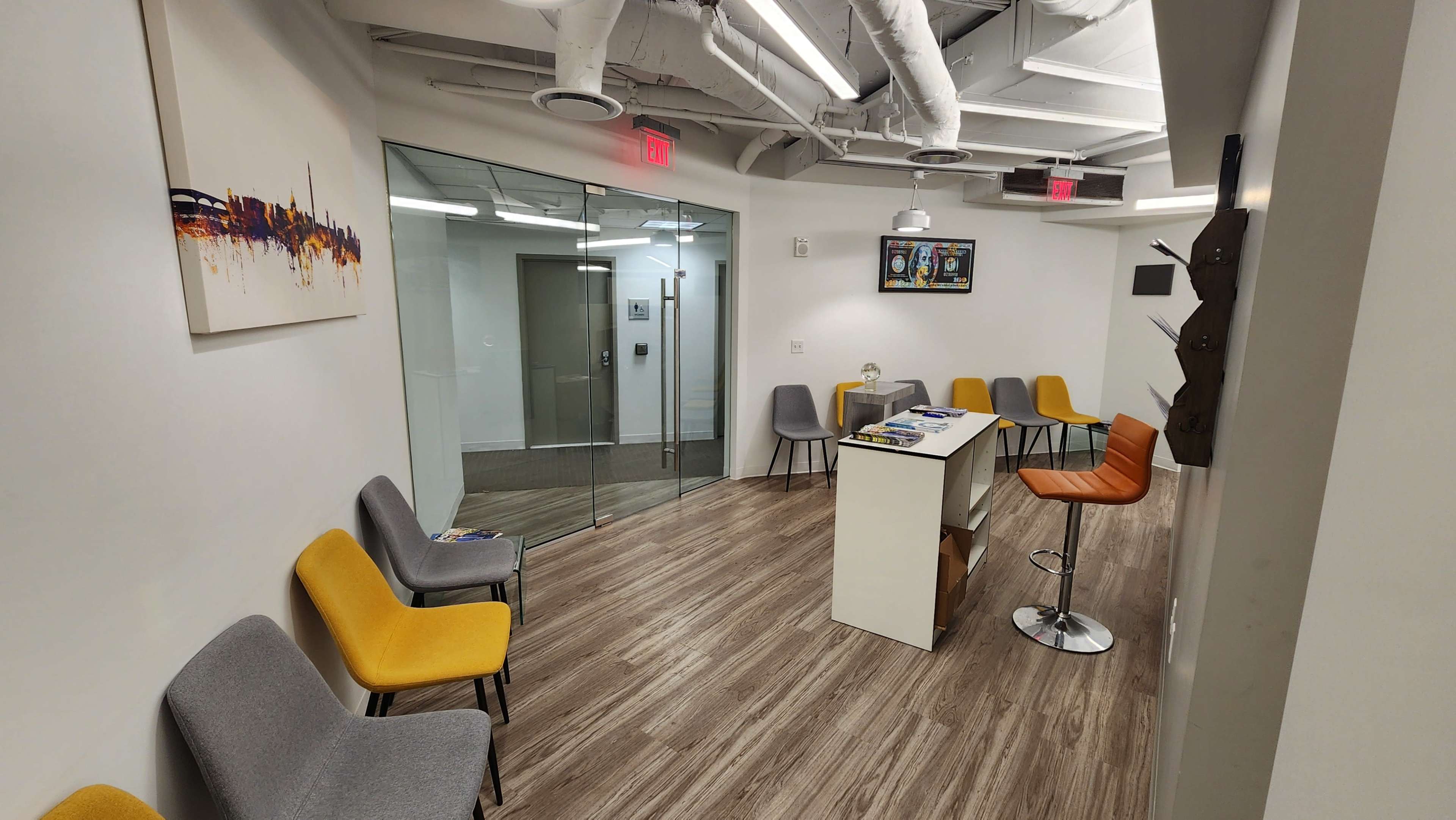 A modern waiting area with a central table surrounded by colorful chairs and a glass partition leading to another room.