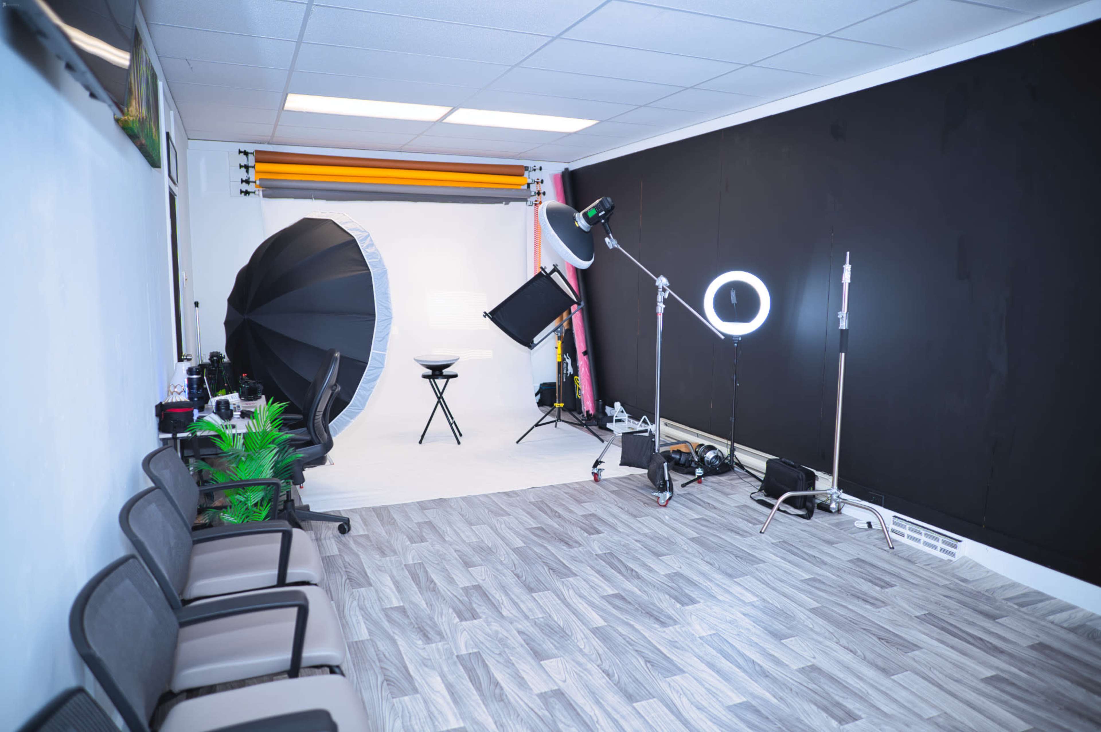 A photography studio with a backdrop setup, lighting equipment, a stool, and several chairs arranged along the wall.