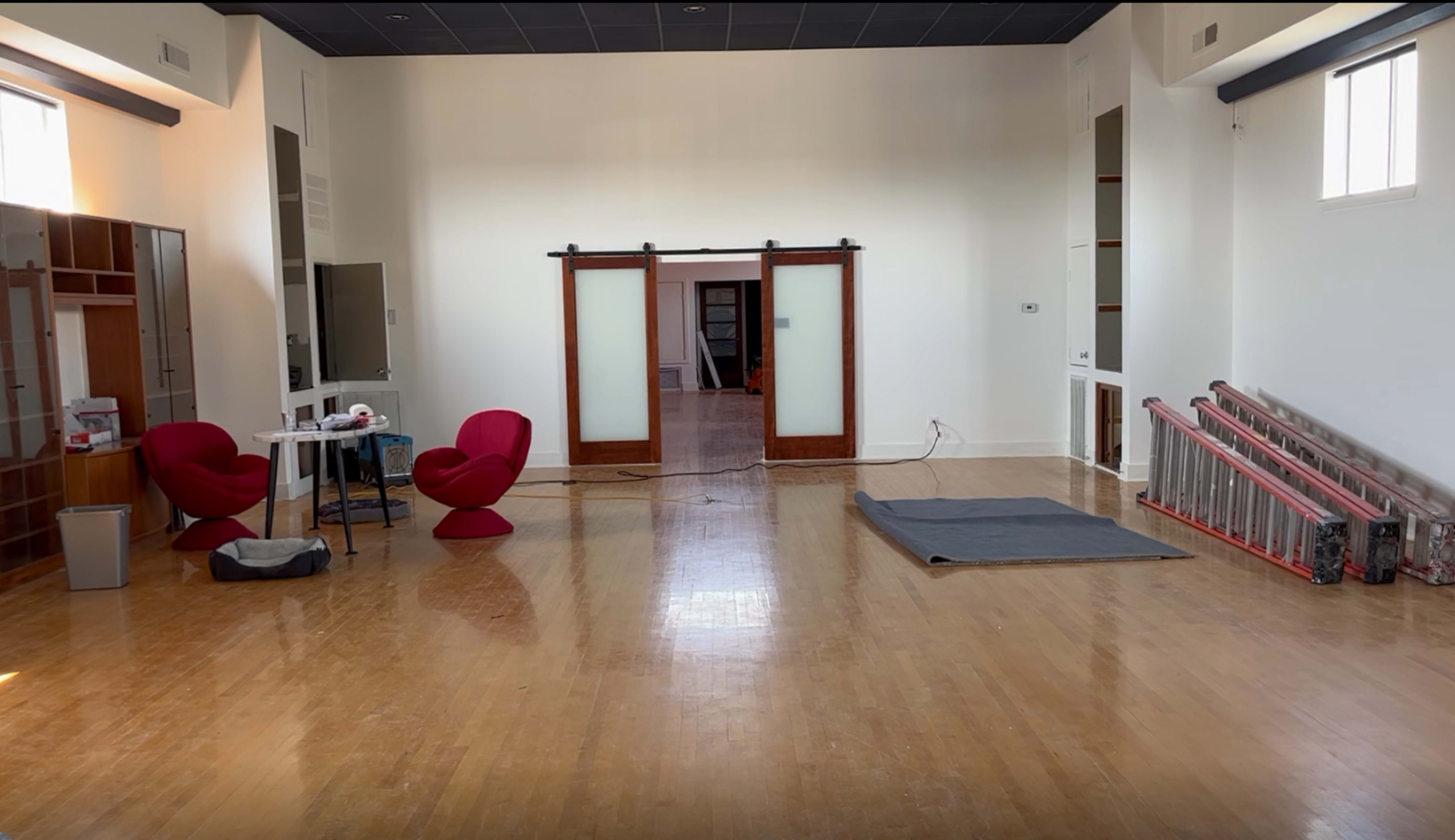 A spacious, empty room with wooden floors, featuring two red chairs, a small table, and a gray mat on the floor.