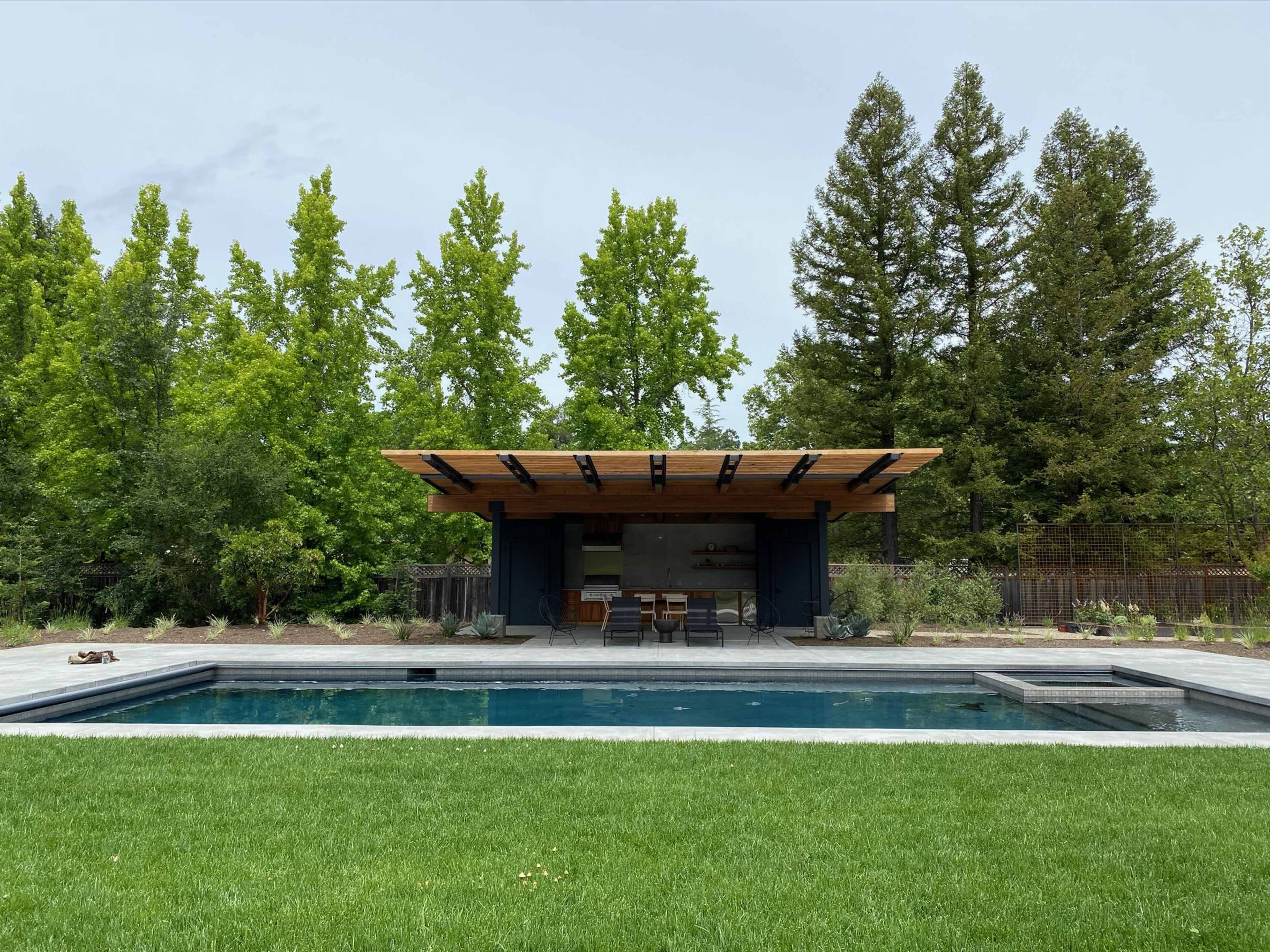 A modern pool house with a wooden roof overlooks a rectangular swimming pool surrounded by a landscaped area of grass and trees.