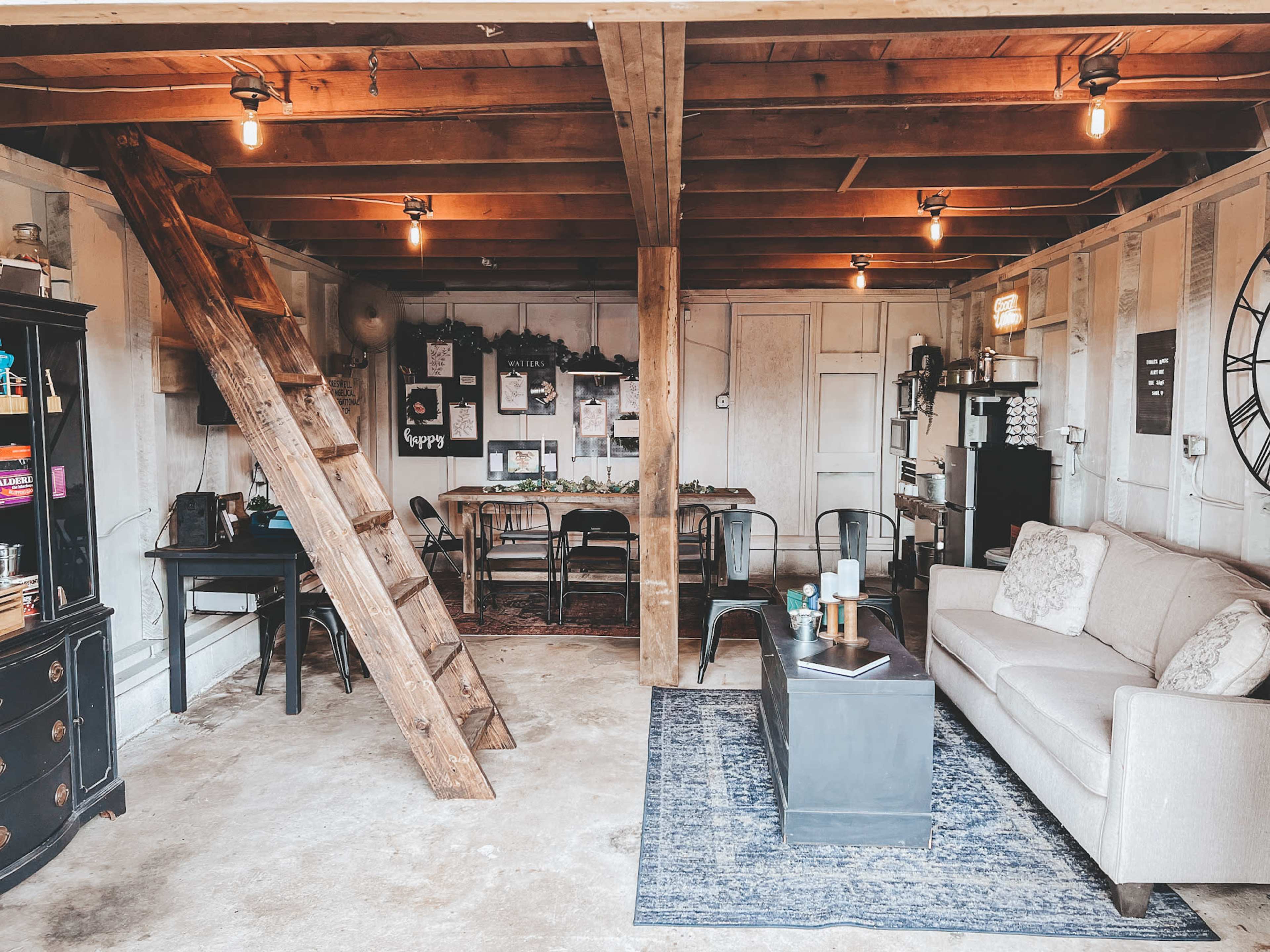 The image shows a garage interior converted into a cozy living space featuring a sofa, a table with chairs, a ladder leading to an upper area, and various decorative elements on the walls.