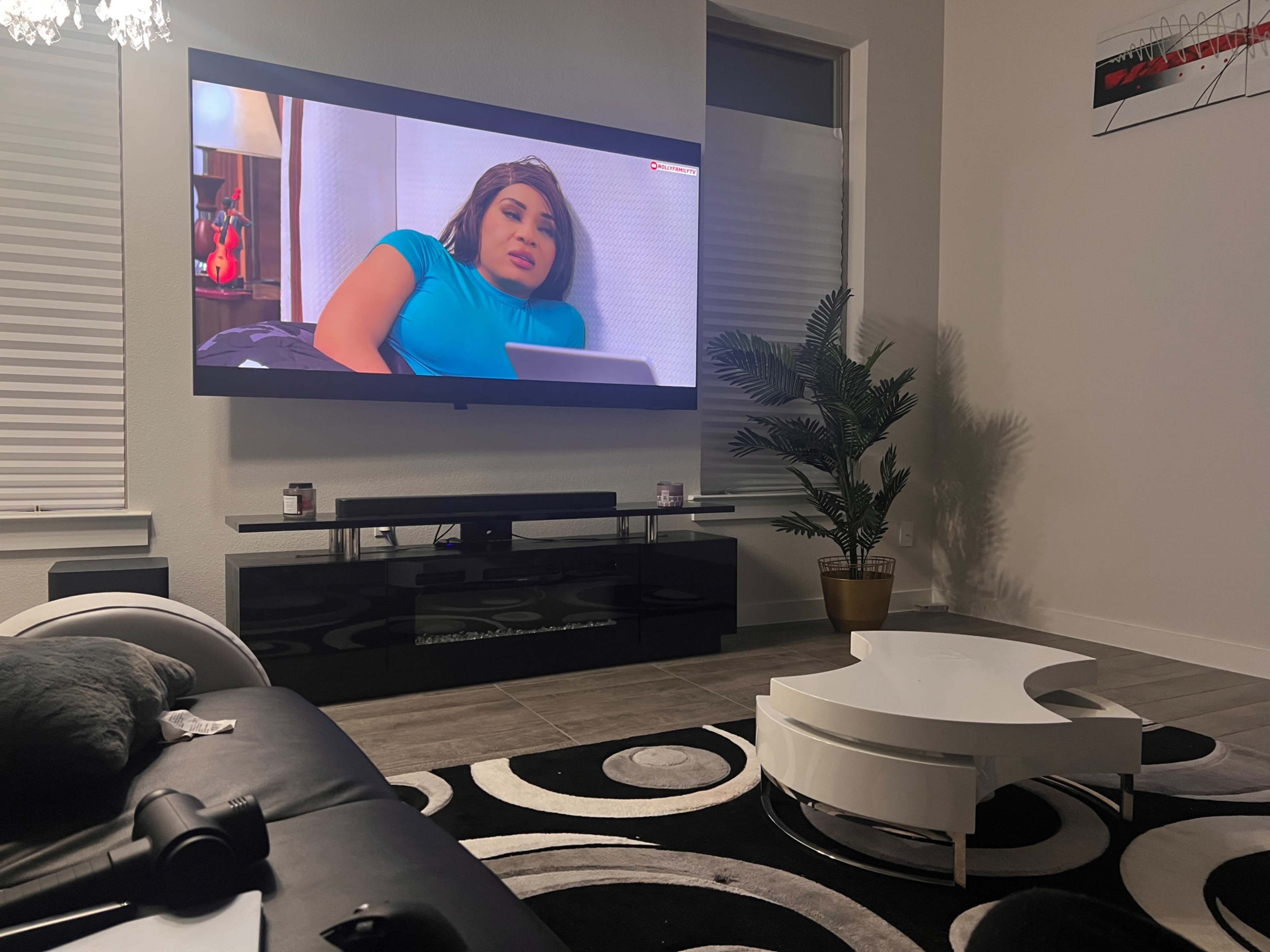 A person is reclining on a sofa while watching a television that displays a scene from a show in a modern living room with a plant and a coffee table.