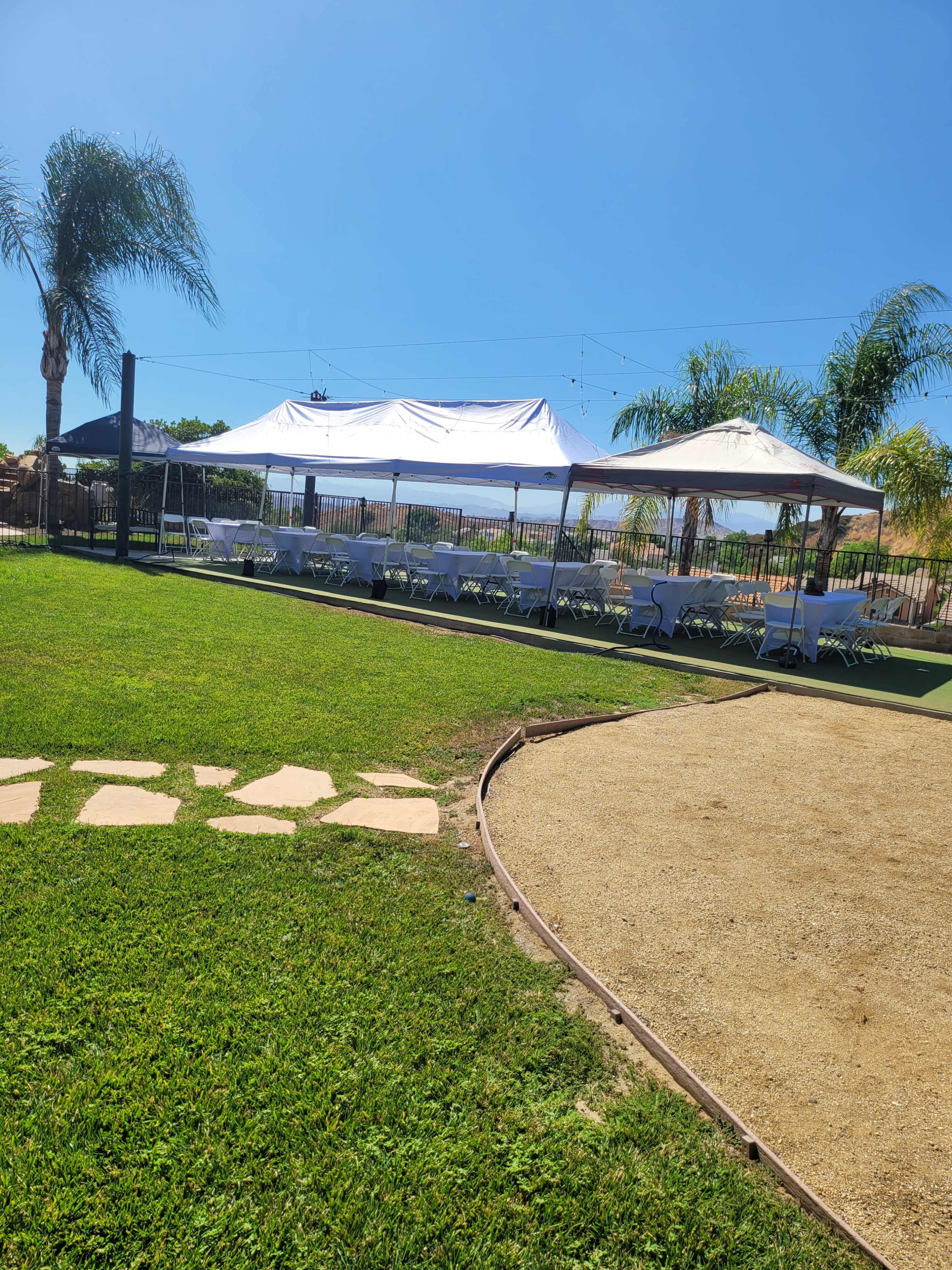 A large white tent with tables and chairs is set up on green grass near a sandy area, surrounded by palm trees against a clear blue sky.