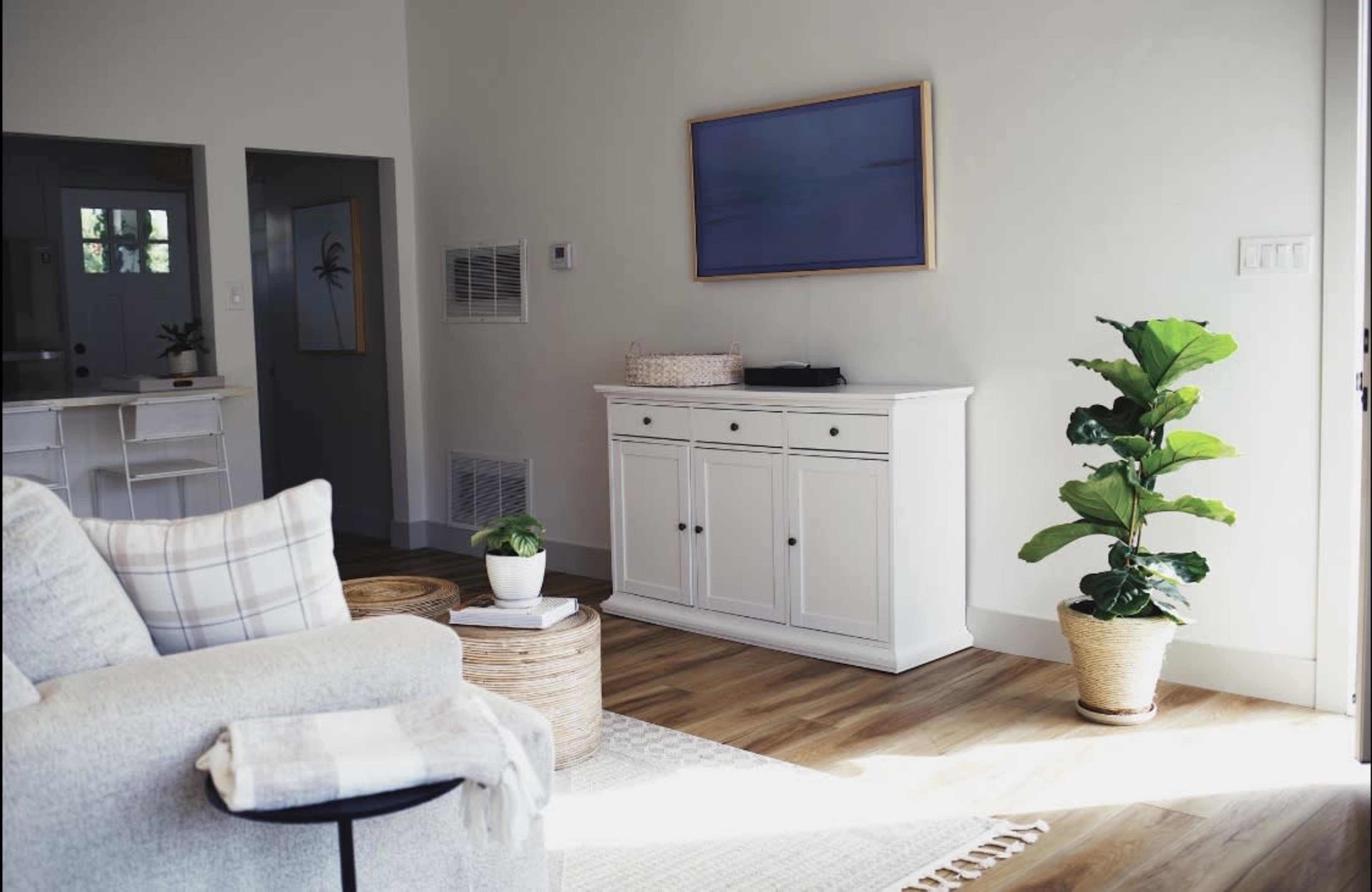 The image shows a bright living room featuring a white cabinet, a potted plant, and a comfortable sofa with a plaid throw, all set on wooden flooring.