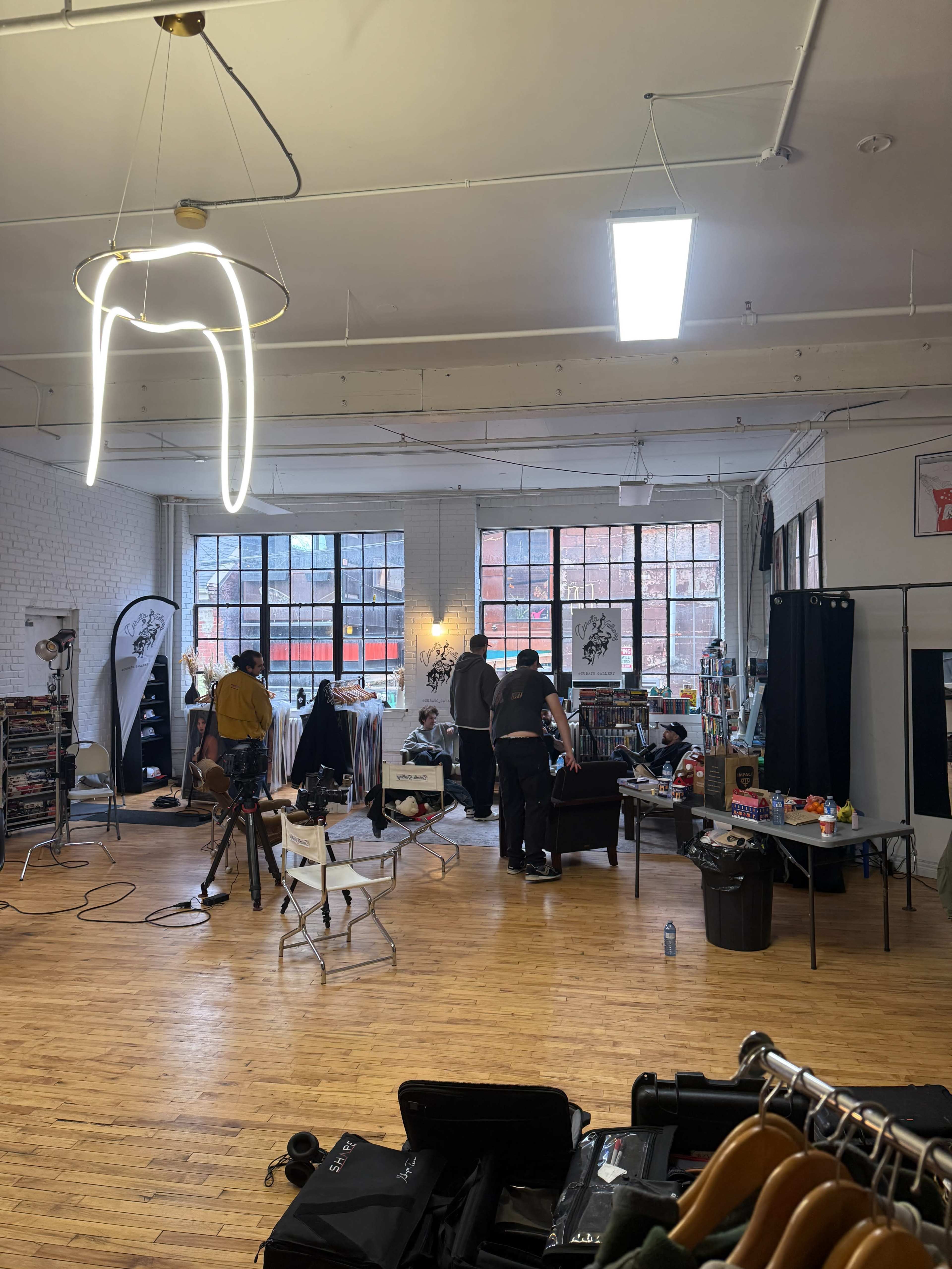 The image shows a spacious studio interior with large windows, a few people working on a project, and various equipment and clothing items scattered throughout the room.