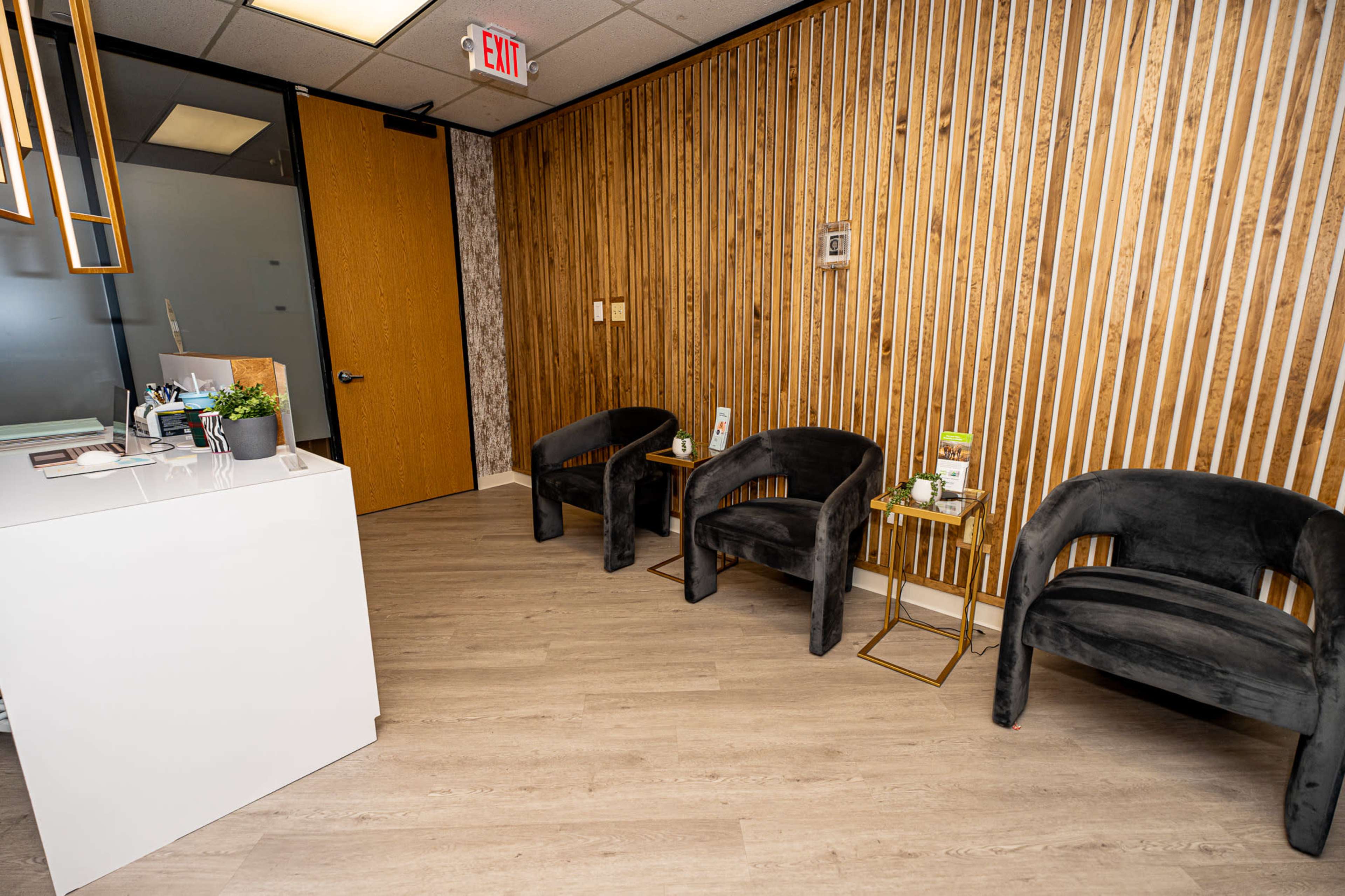 The image shows a modern waiting area with three black chairs against a wooden slat wall and a white reception desk.