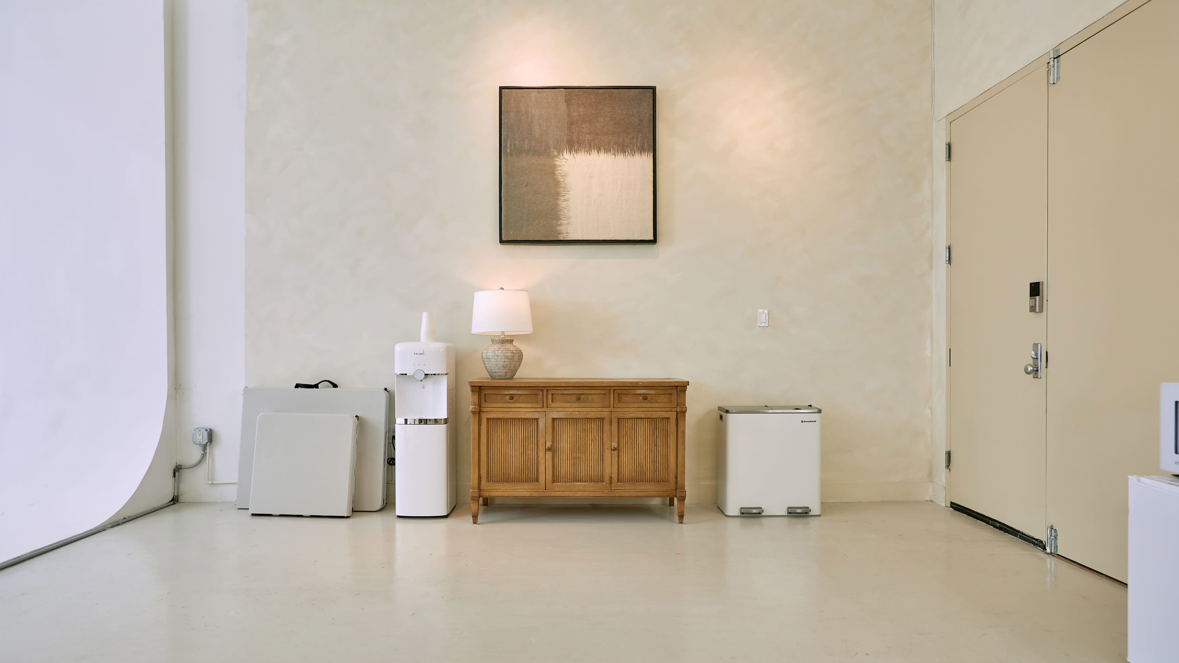 The image shows a minimalist room with light-colored walls, featuring a wooden cabinet, a lamp, a small refrigerator, a water dispenser, and a door.