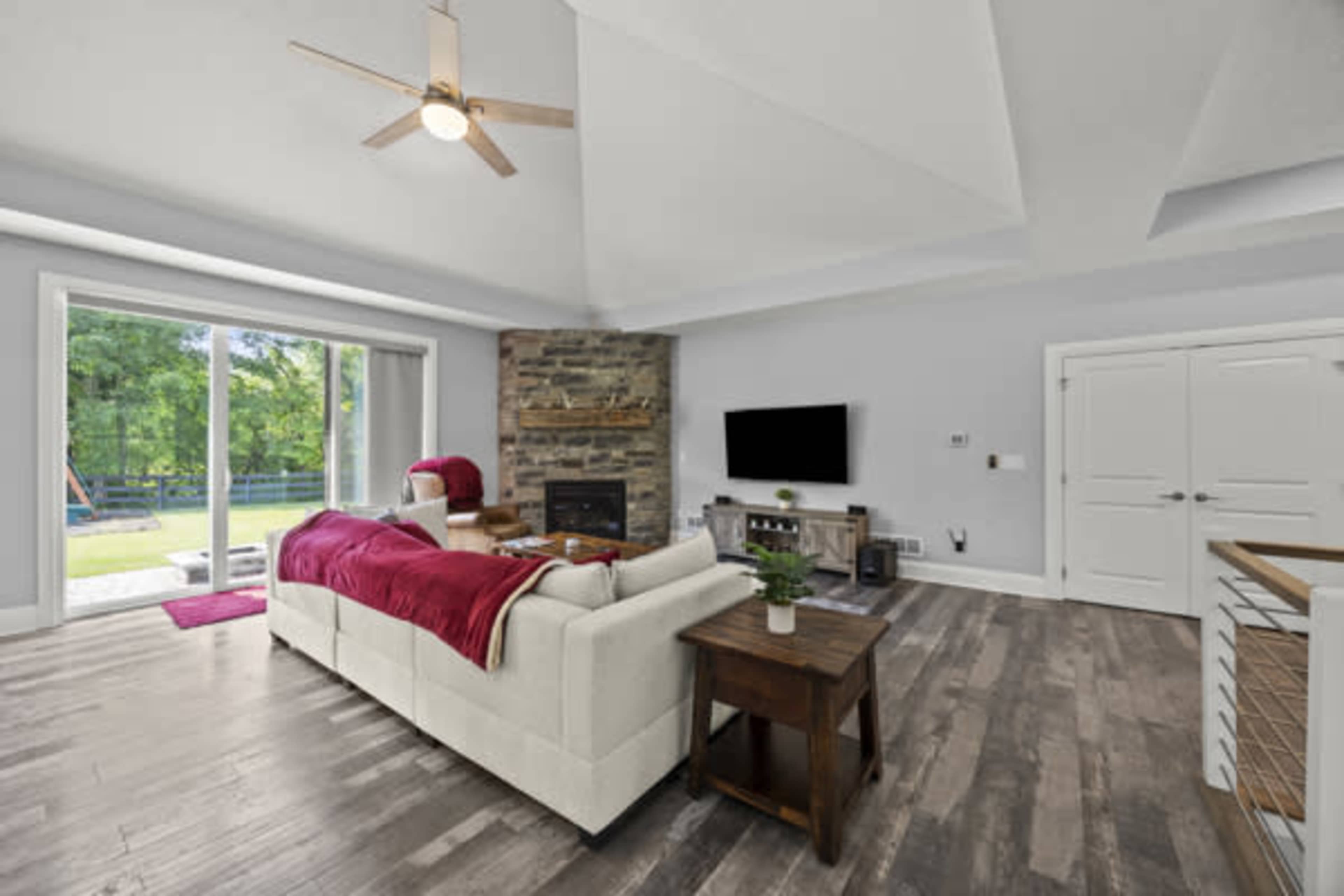 The image shows a modern living room featuring a beige sofa, a stone fireplace, a wall-mounted television, and large windows that open to an outdoor view.