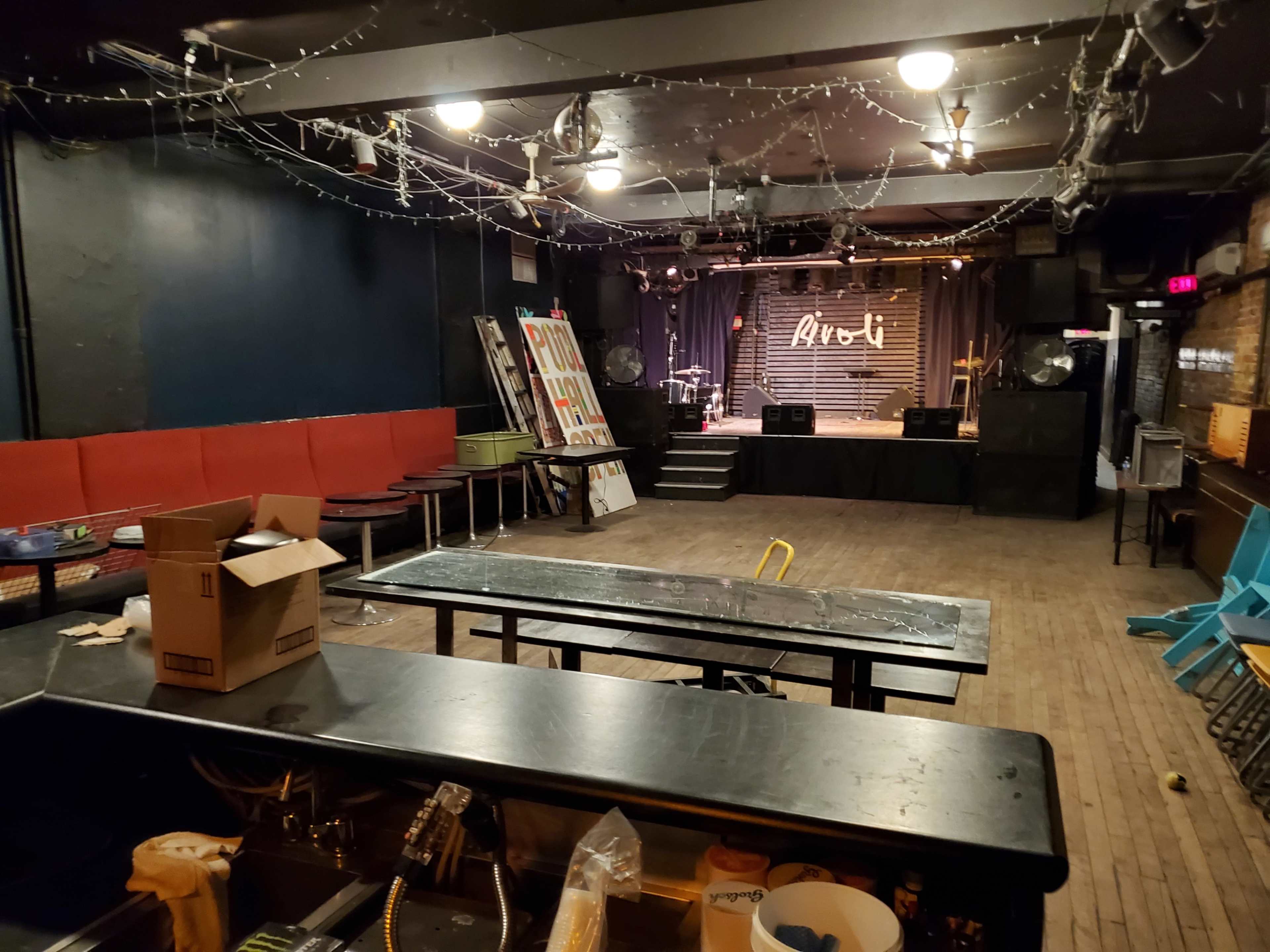 The image shows an empty performance venue with a stage, a bar counter, and red seating areas.