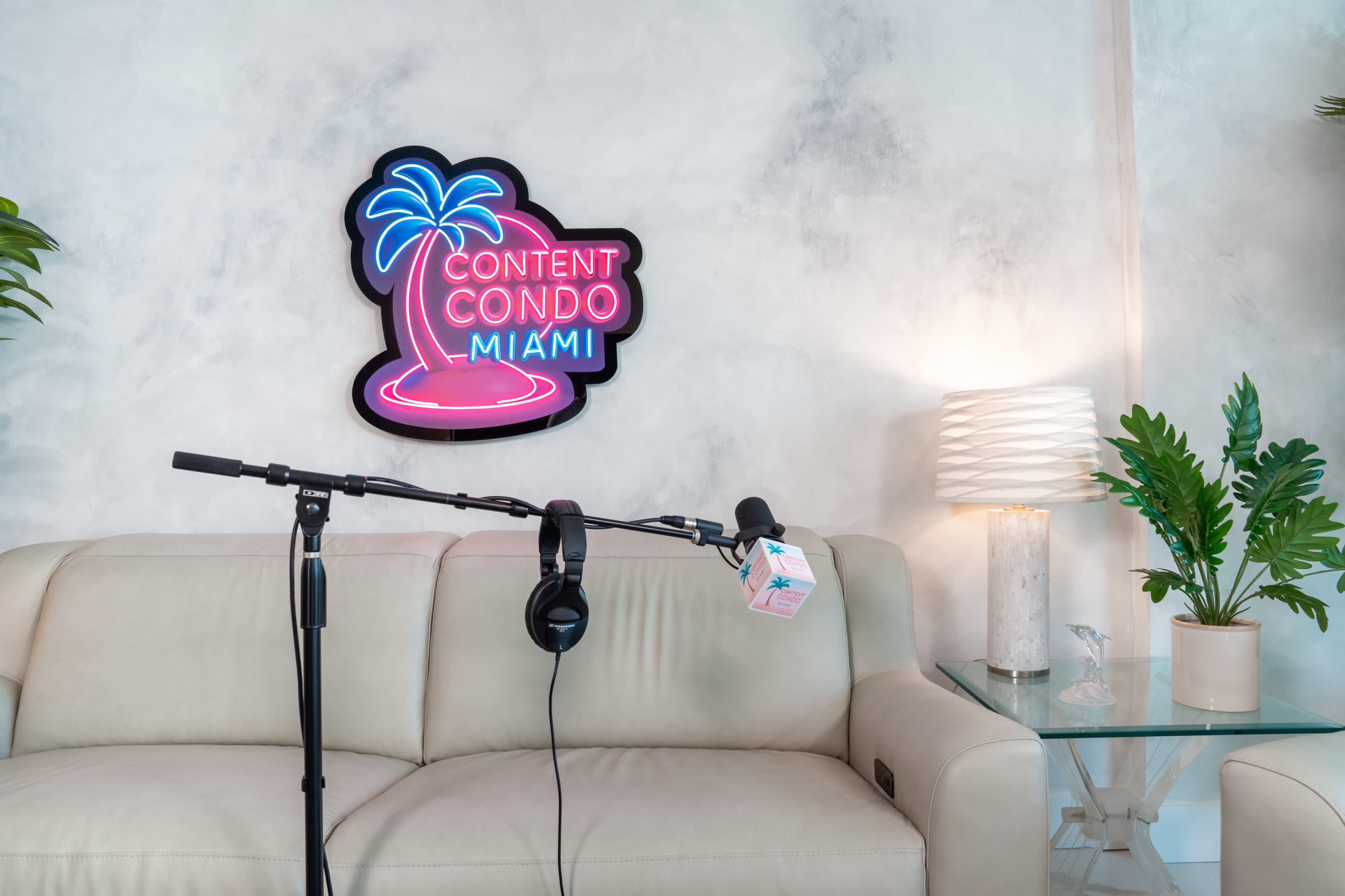 The image shows a modern lounge area with a beige sofa, a microphone on a stand, headphones, a neon sign that says "CONTENT CONDO MIAMI," and a lamp next to a glass side table with a plant.