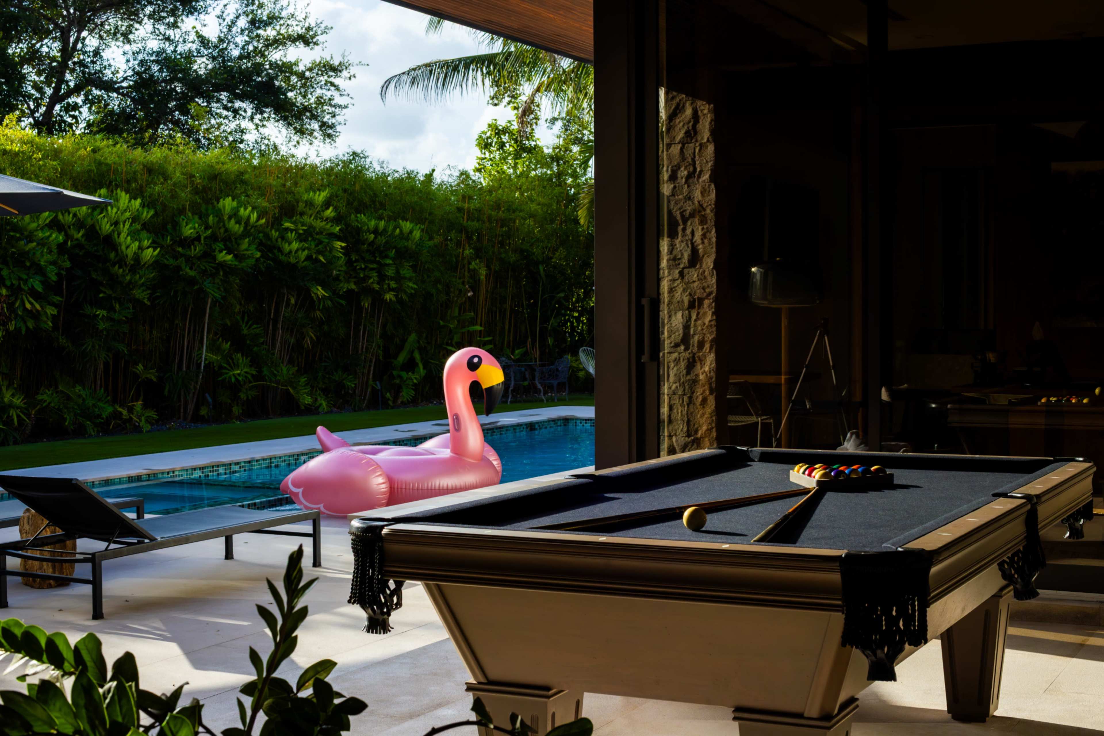 A pink inflatable flamingo floats in a swimming pool visible from an indoor pool table area surrounded by lush greenery.