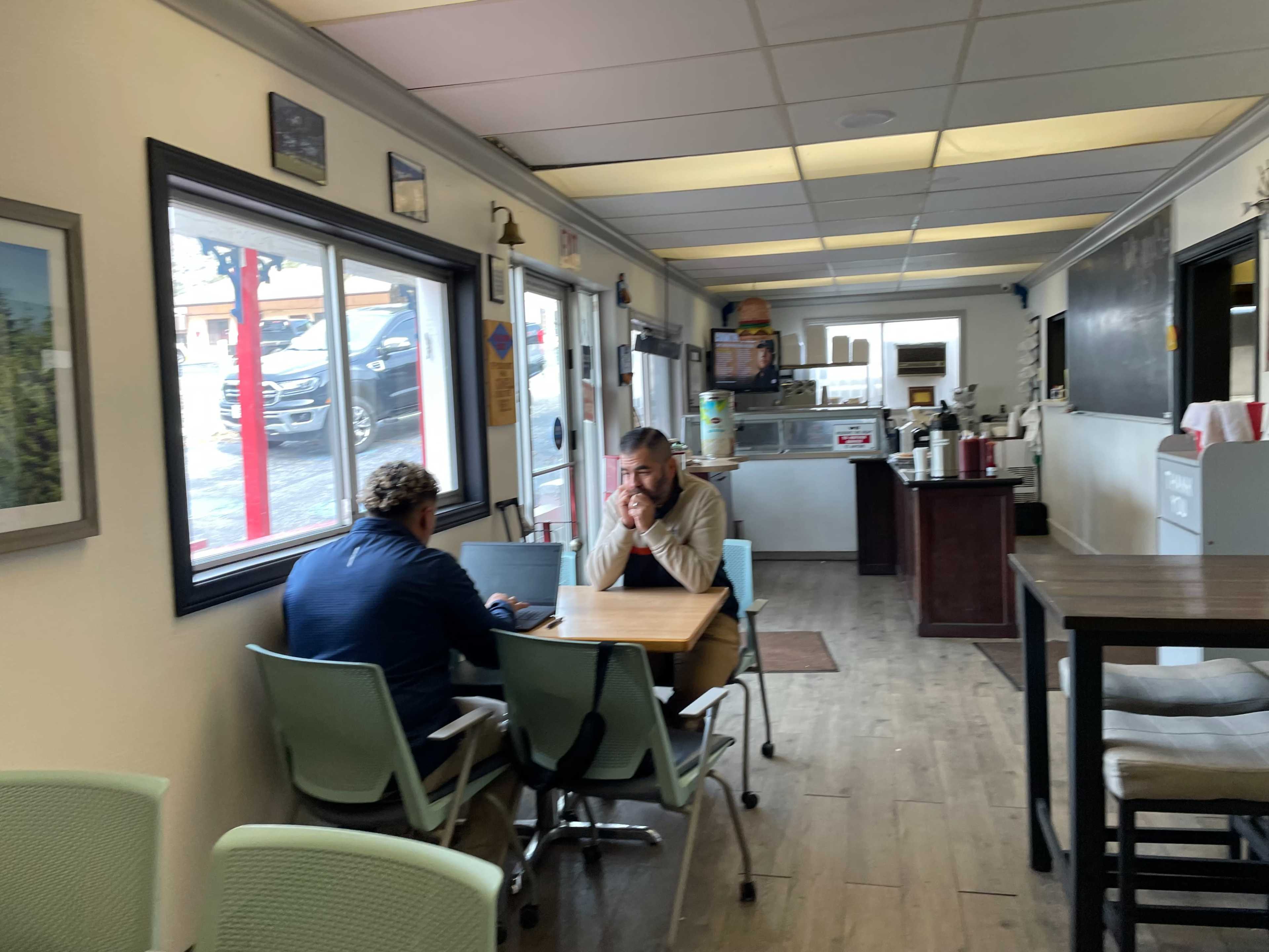 Two men sitting at a table in a casual dining area, with a kitchenette visible in the background.