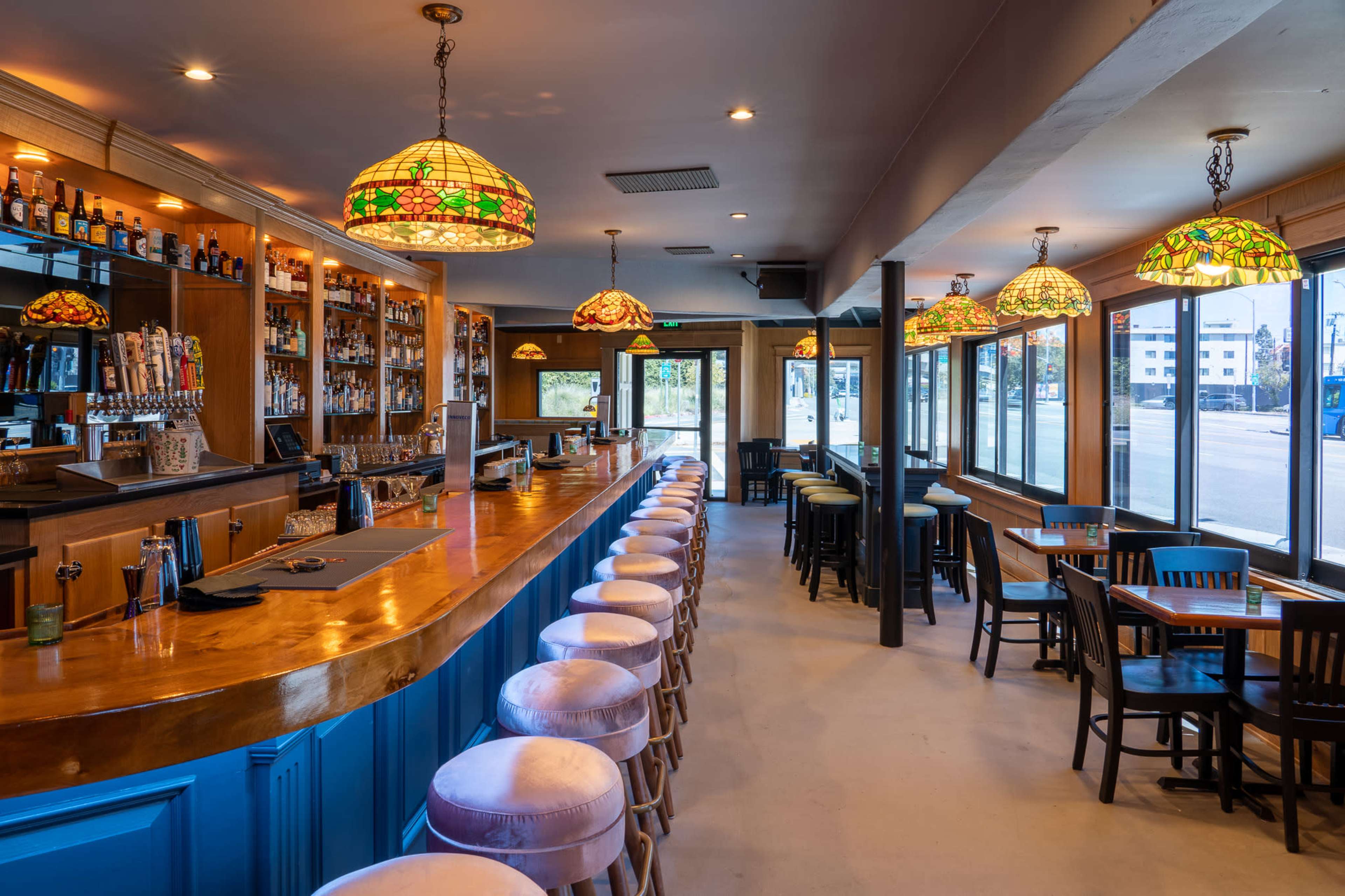 A well-lit bar area with a wooden counter, high stools, and stained glass pendant lights, adjacent to large windows facing the street.