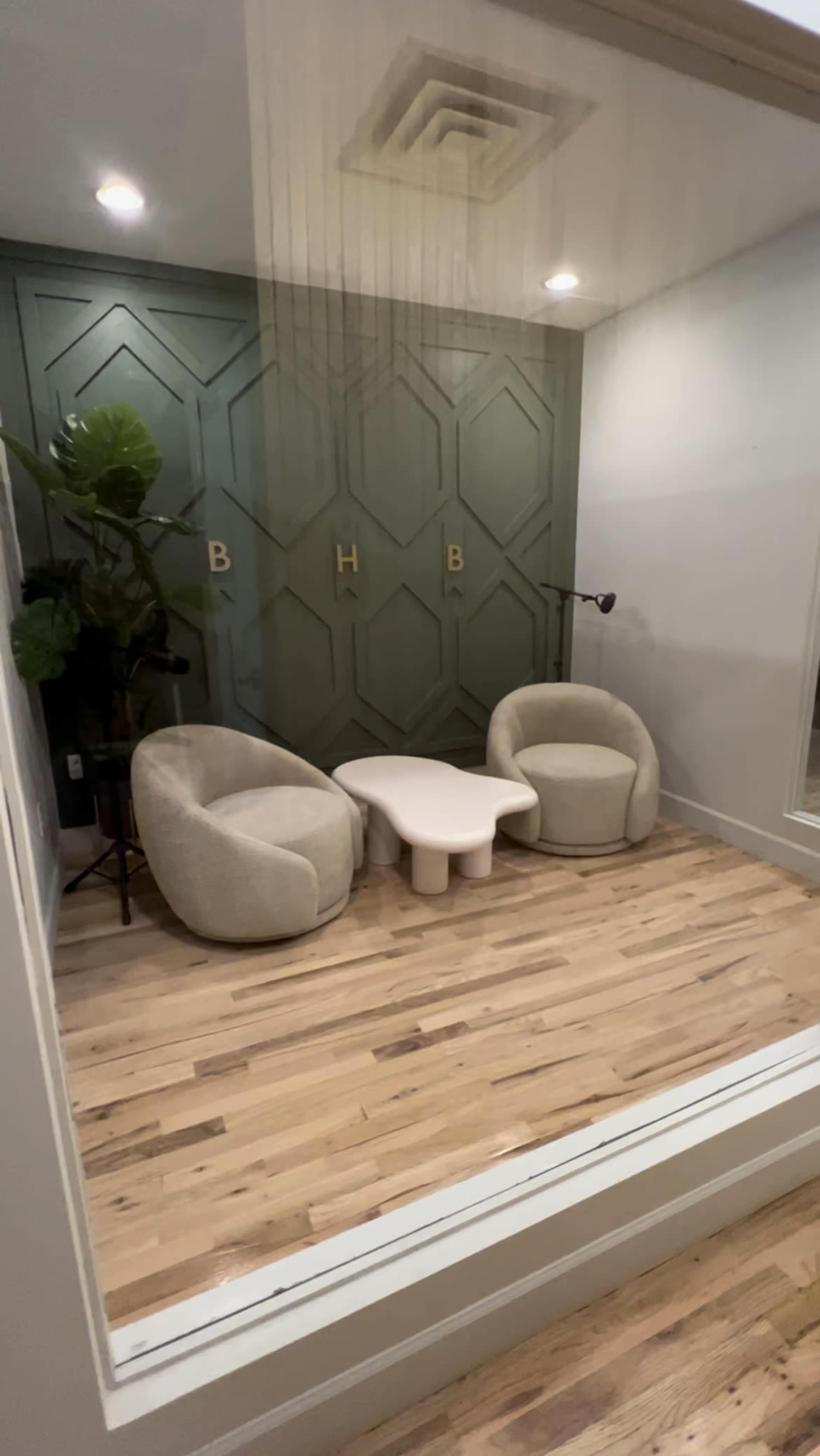 A small interior space features two cream-colored chairs and a light-colored coffee table on wooden flooring, with a green geometric-patterned accent wall and a potted plant.