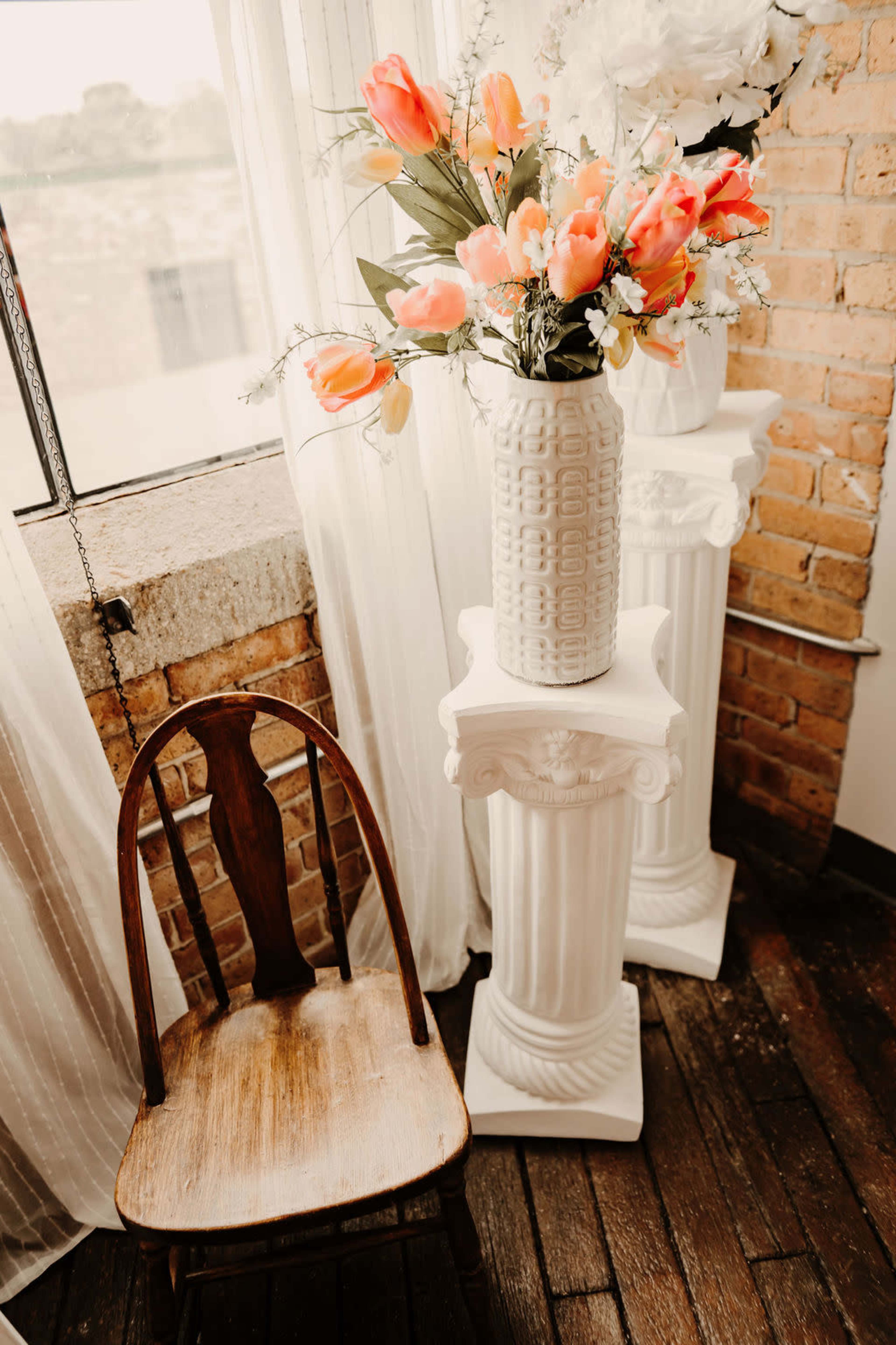 A wooden chair sits next to two white pedestals, each topped with a vase of orange flowers, in a room with a brick wall and large window.