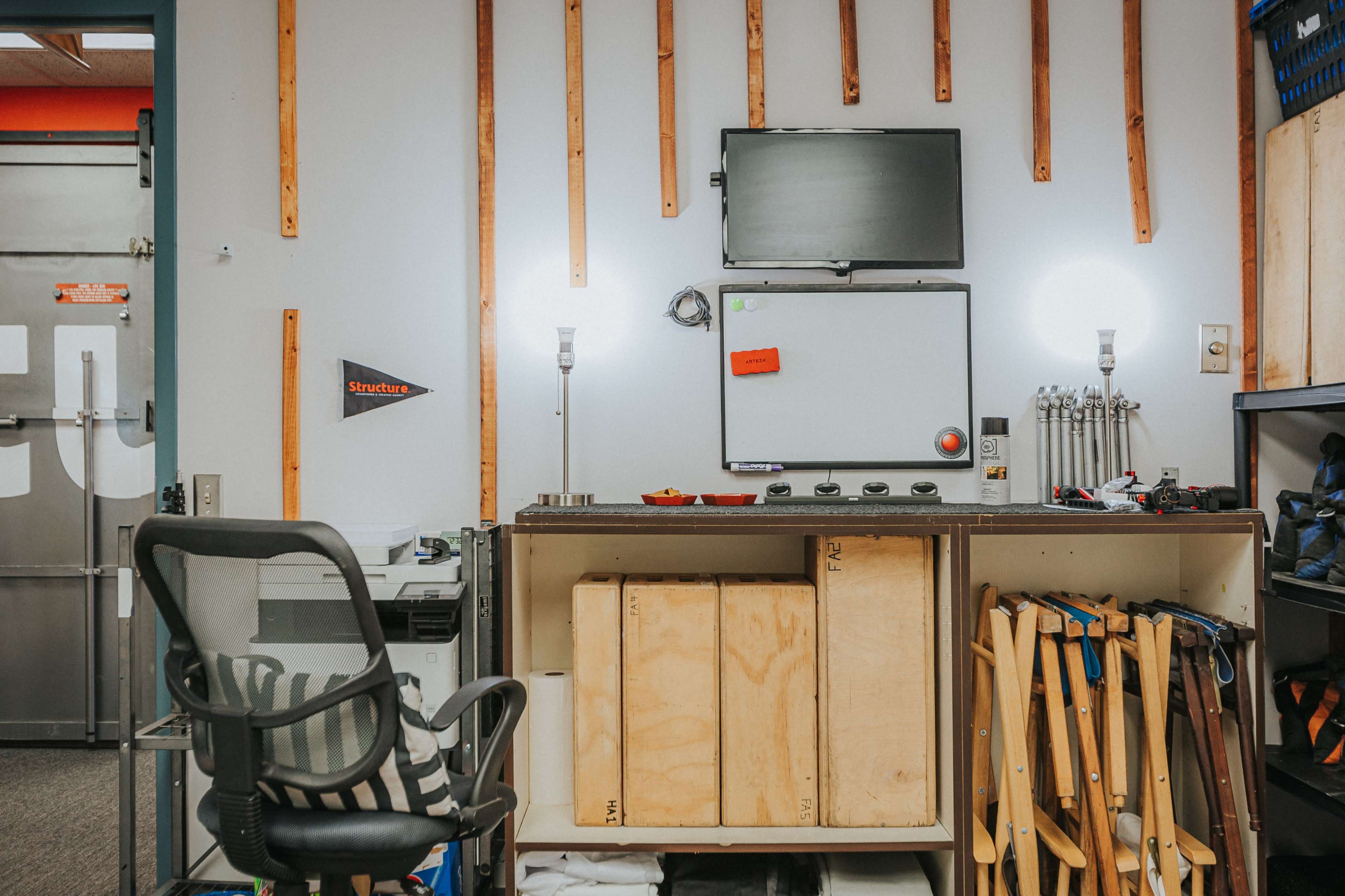 A workshop with a desk area featuring a monitor, whiteboard, and shelving containing tools and equipment.