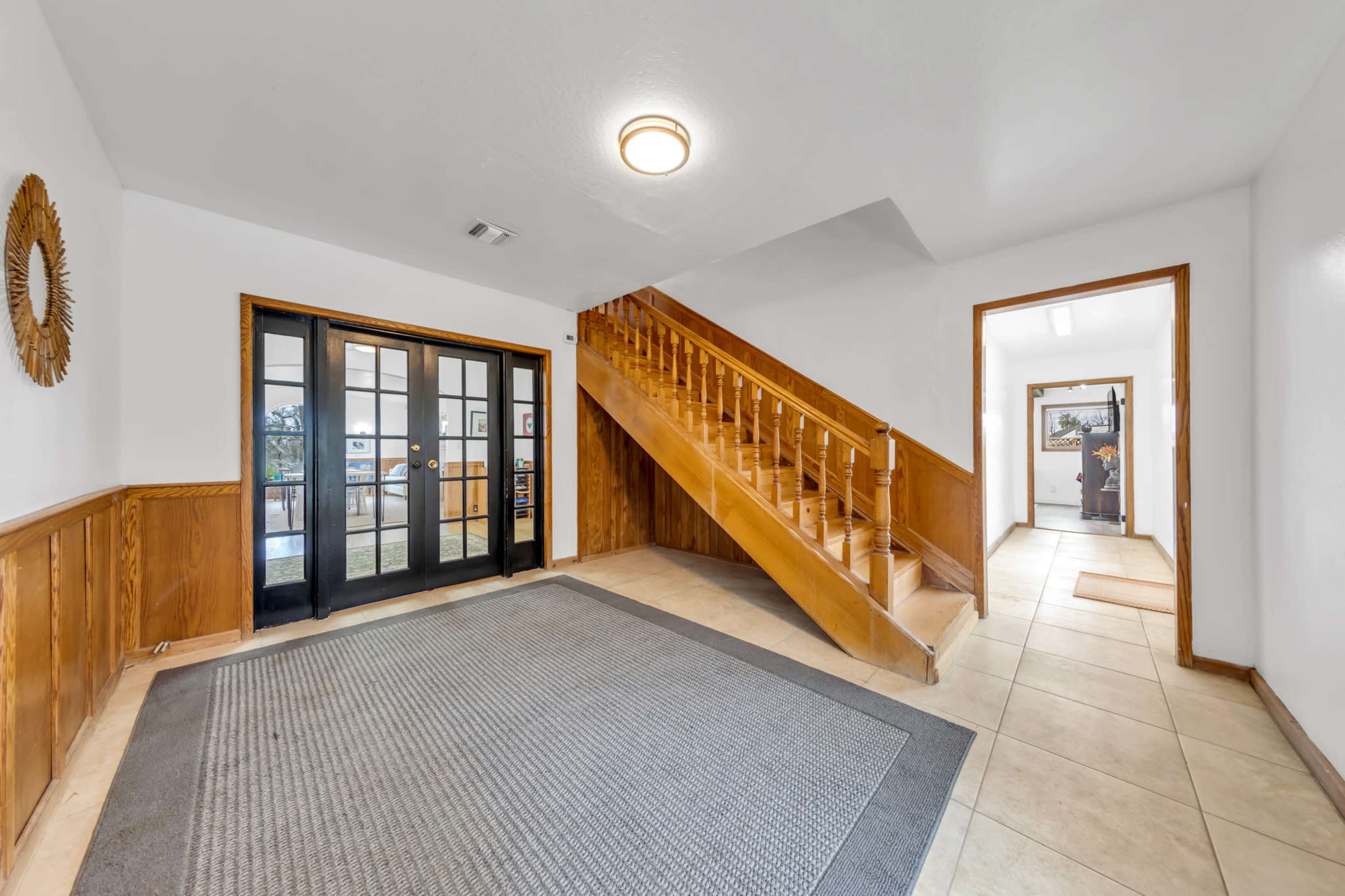 The image shows a spacious entryway with a wooden staircase, ceramic tile flooring, and double glass doors leading to another room.