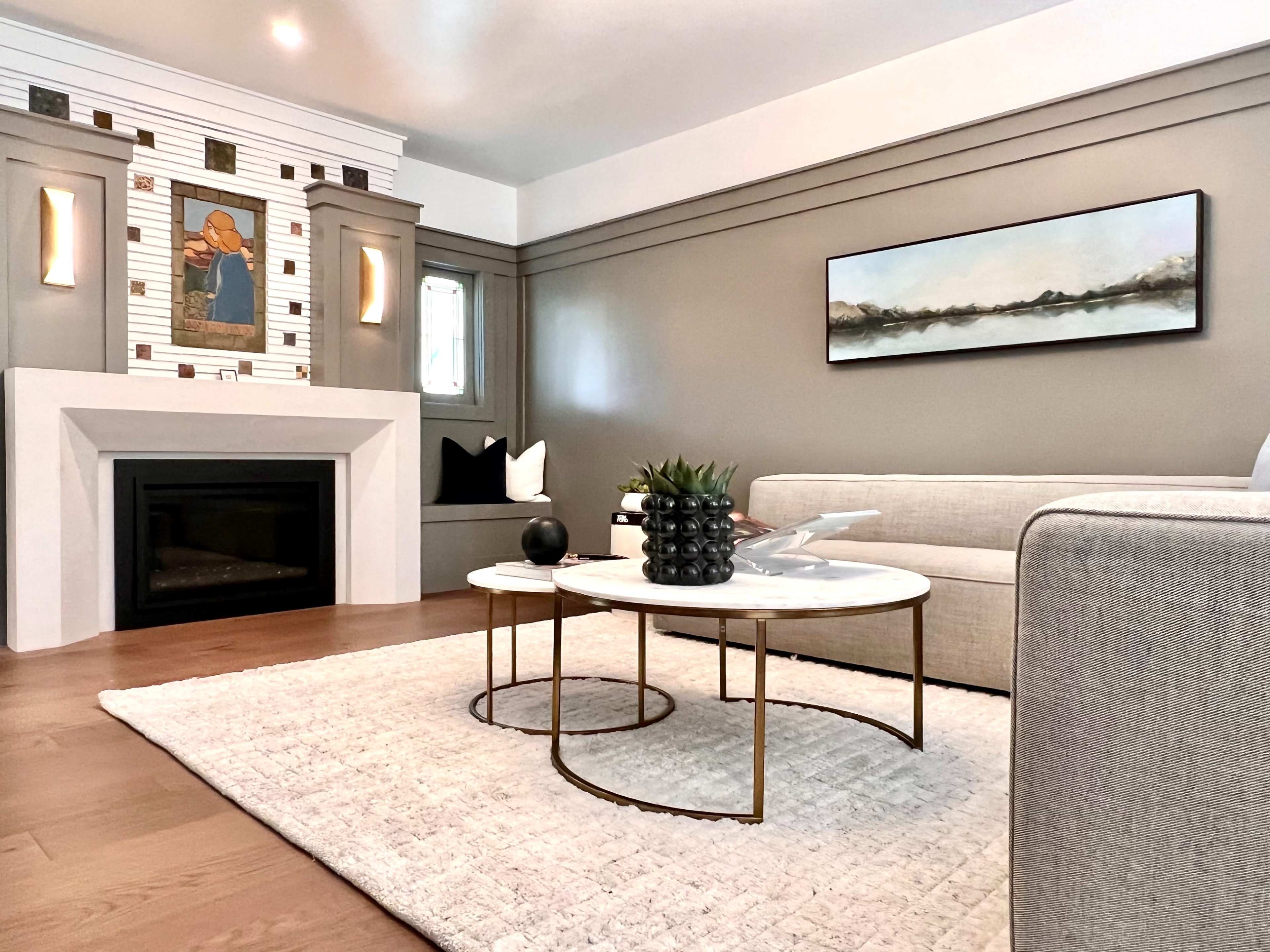A modern living room features a fireplace with artwork above it, a beige sofa, two round coffee tables, and a light area rug.