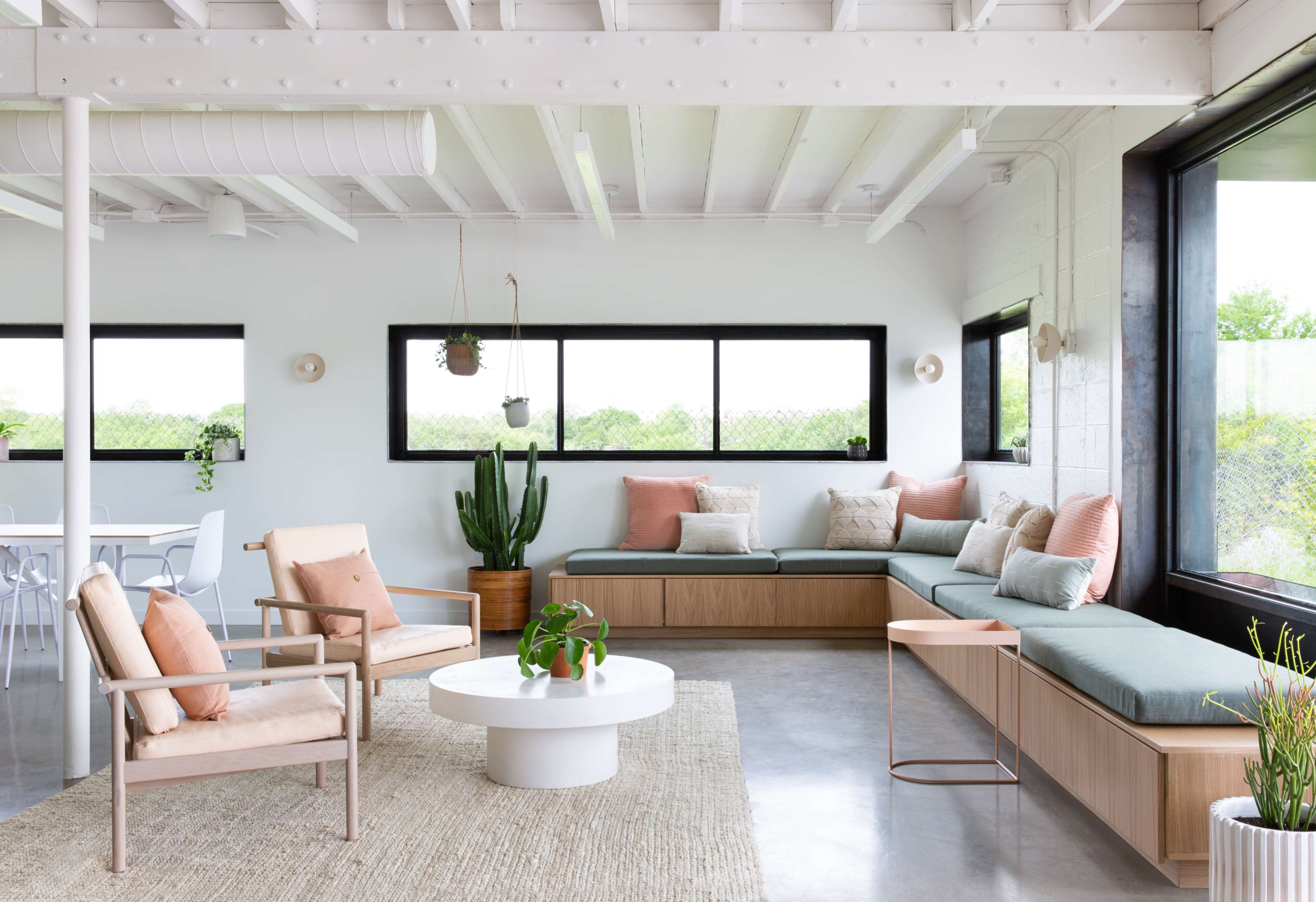 A modern living space with a light color scheme, featuring a sectional seating area with cushions, a round coffee table, and large windows that provide a view of outdoor greenery.