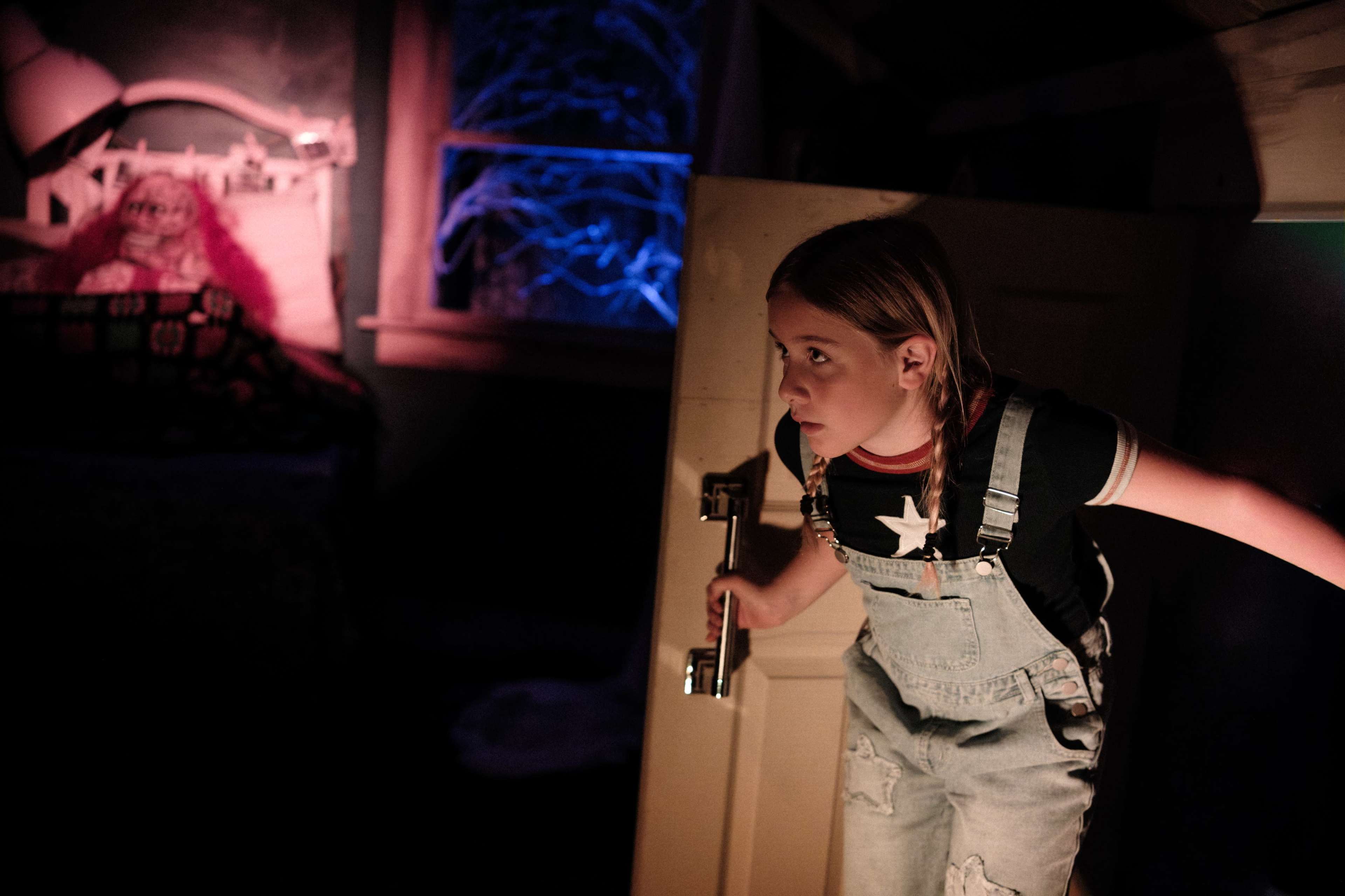 A young girl with blonde hair wearing overalls peeks around a partially open door into a dark room, while a doll sits on a bed in the background.