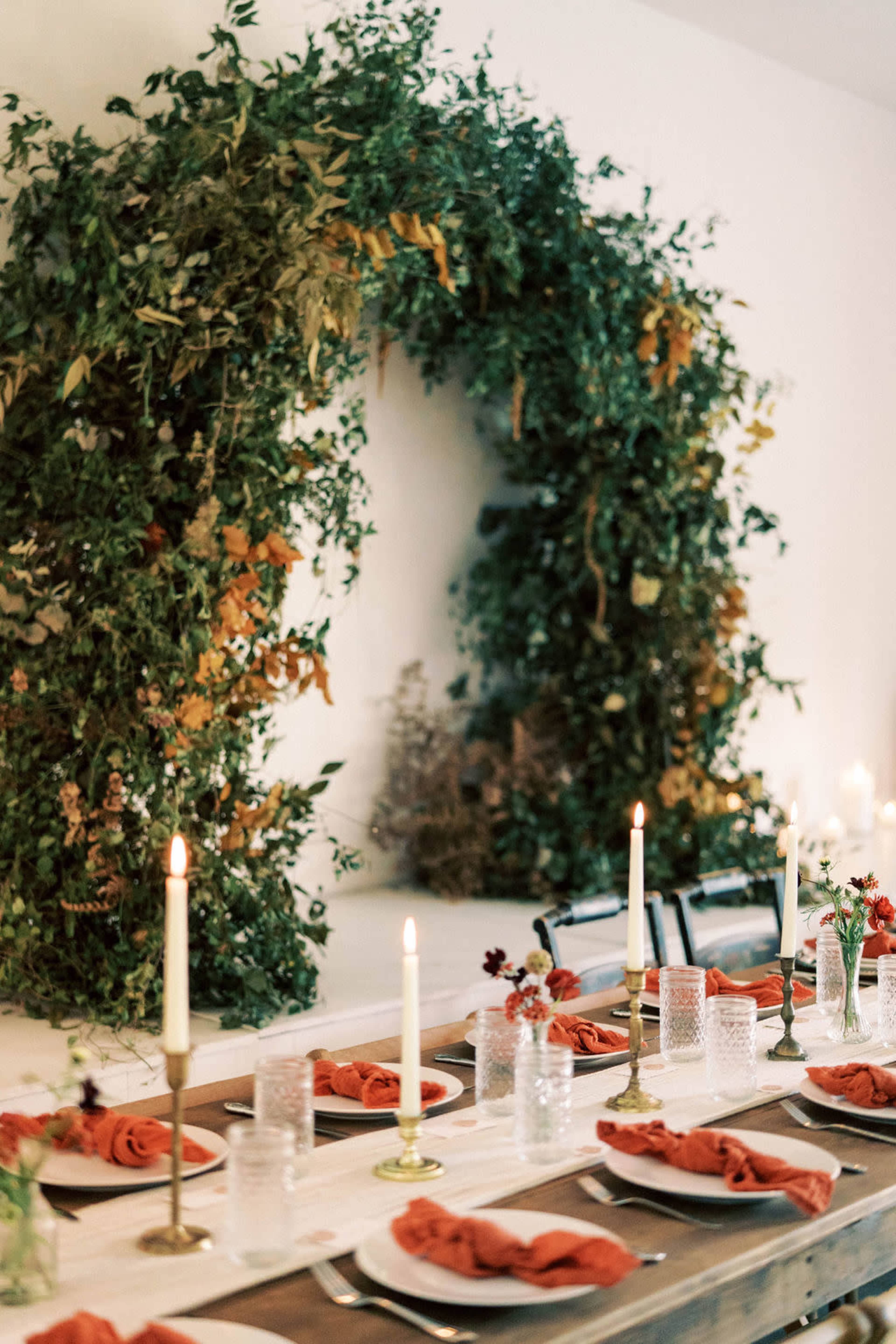 A long dining table is set with red napkins and clear glassware, surrounded by a decorative greenery arrangement on the wall.