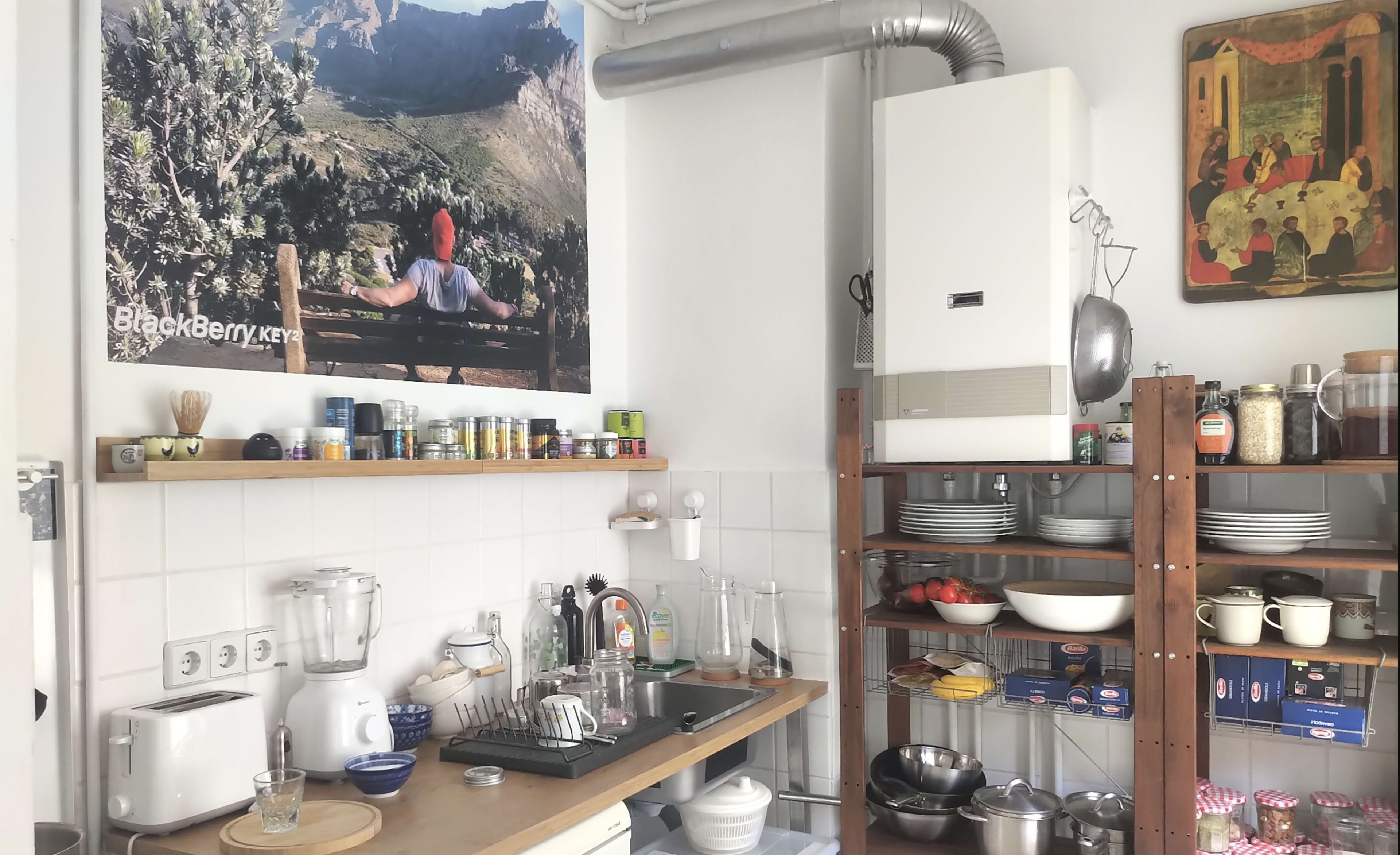 The image shows a cluttered kitchen with a wooden counter, an assortment of dishes, and a large poster on the wall depicting a person overlooking a mountainous landscape.