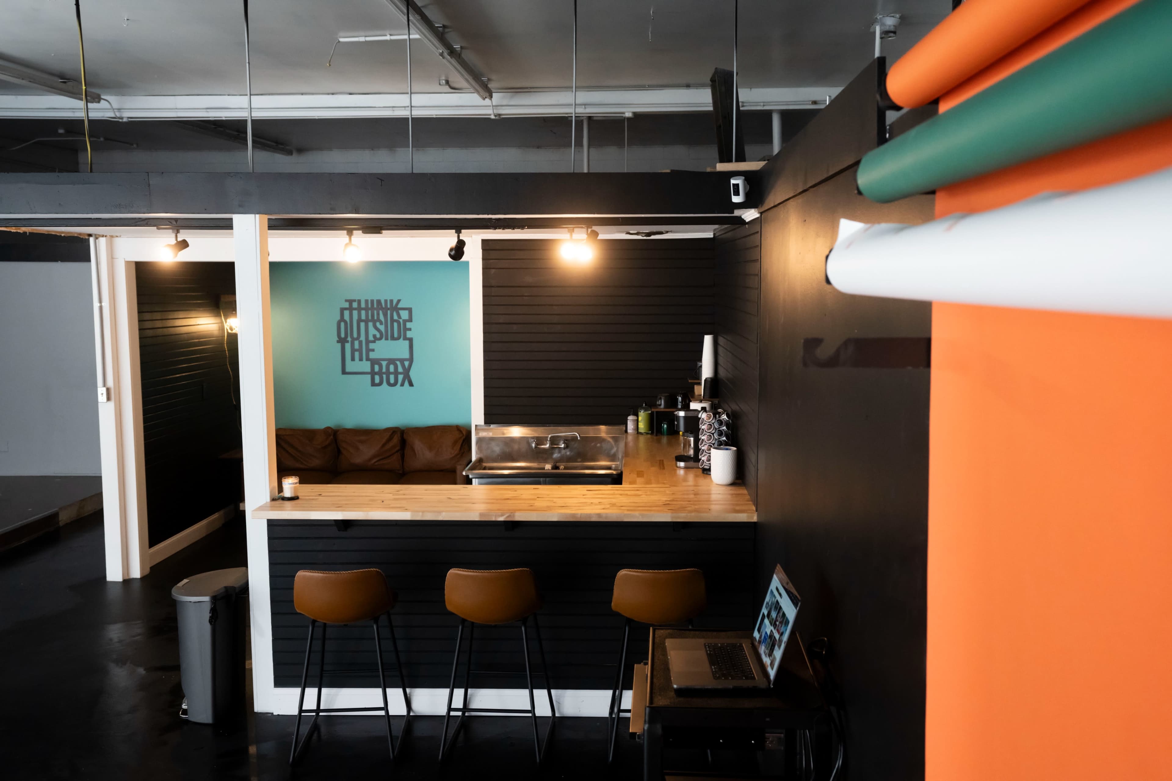 The image shows a modern kitchenette with a wooden counter, three bar stools, a minimalist black wall, and an inspirational quote displayed on the wall.