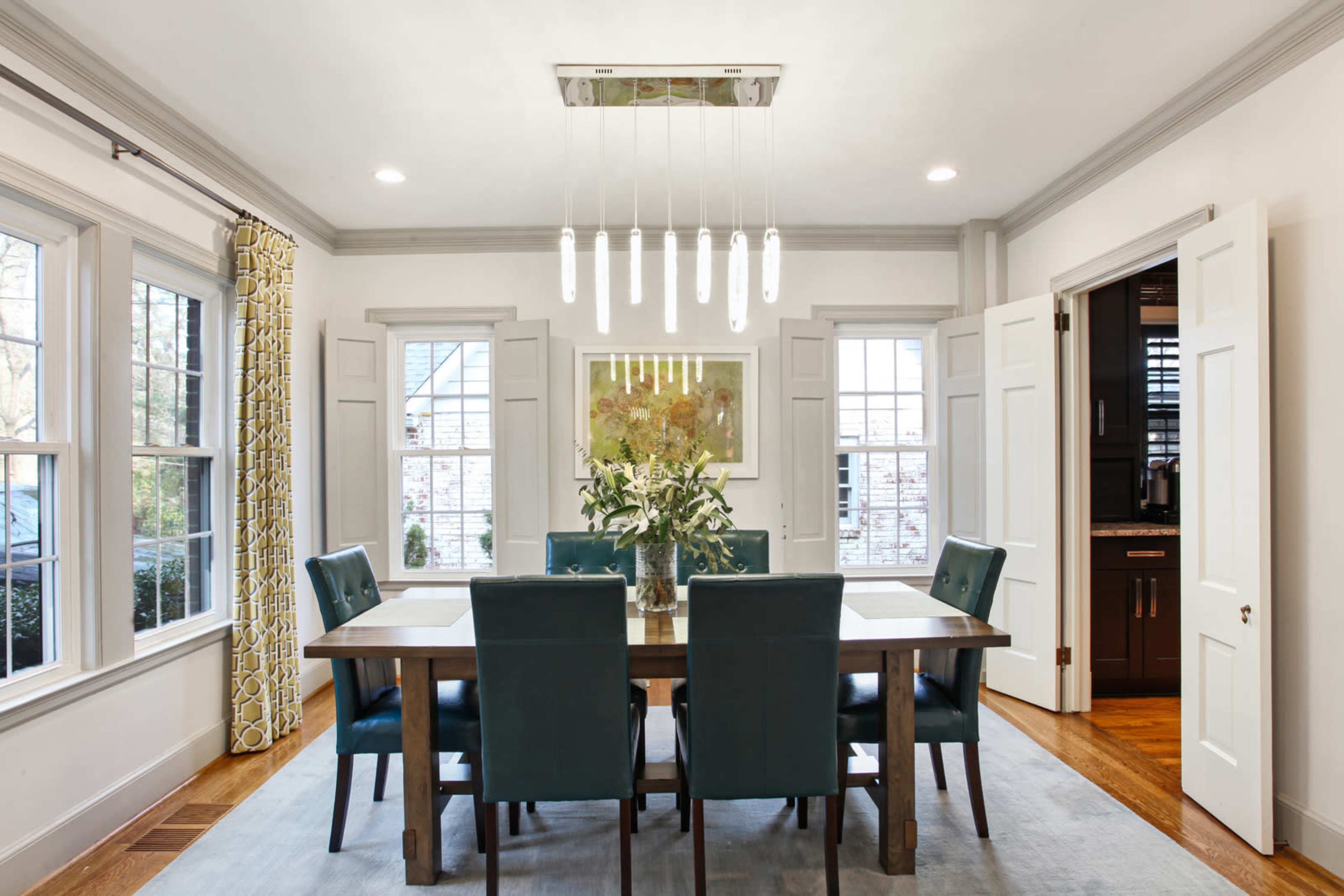 A modern dining room features a rectangular table surrounded by six teal chairs, illuminated by a linear light fixture overhead, with windows flanking the walls.