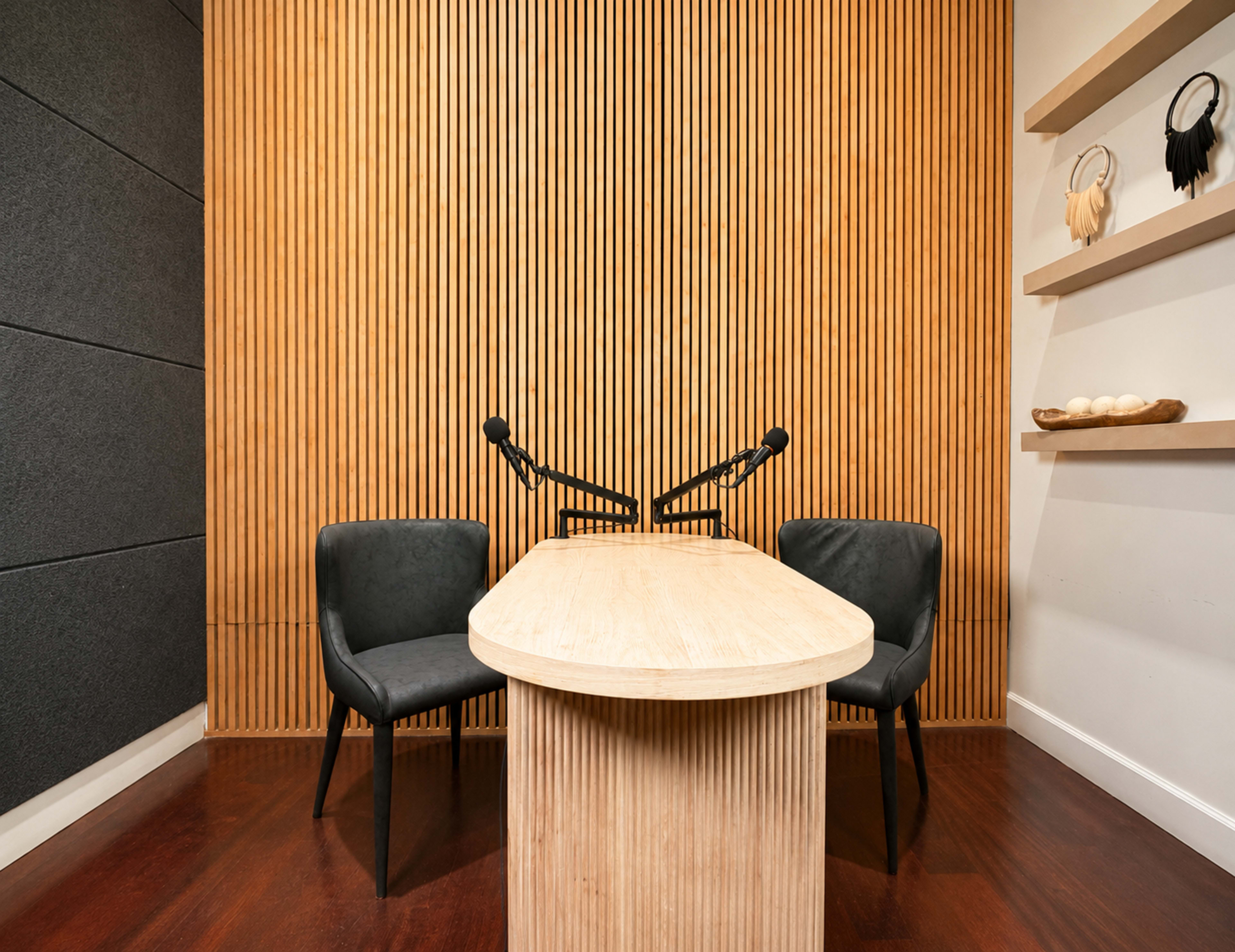A minimalistic podcast recording studio features a rounded wooden table and two black chairs against a backdrop of vertical wooden slats.