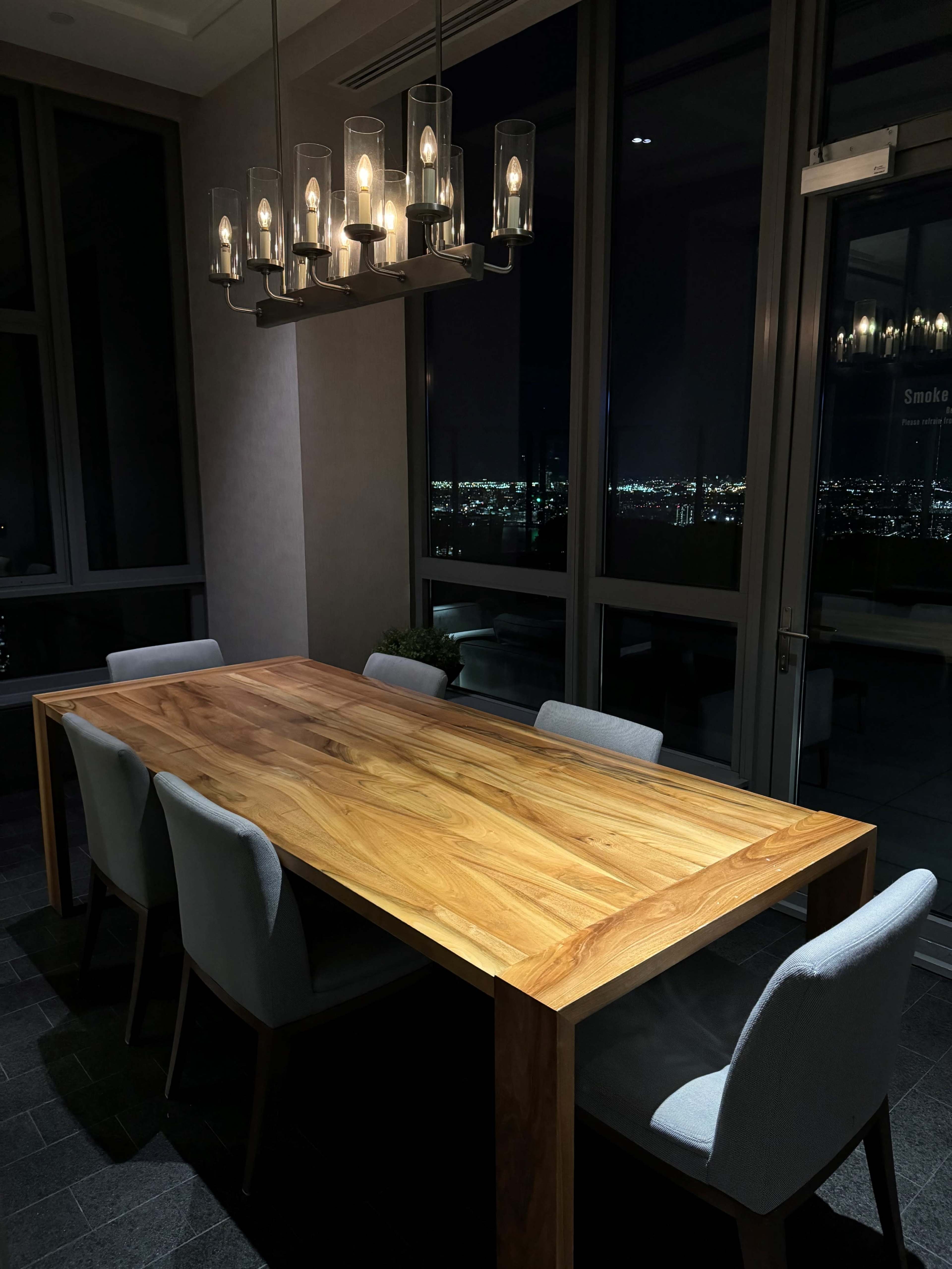A wooden dining table with six upholstered chairs is positioned in a dimly lit room with large windows displaying a city skyline at night.