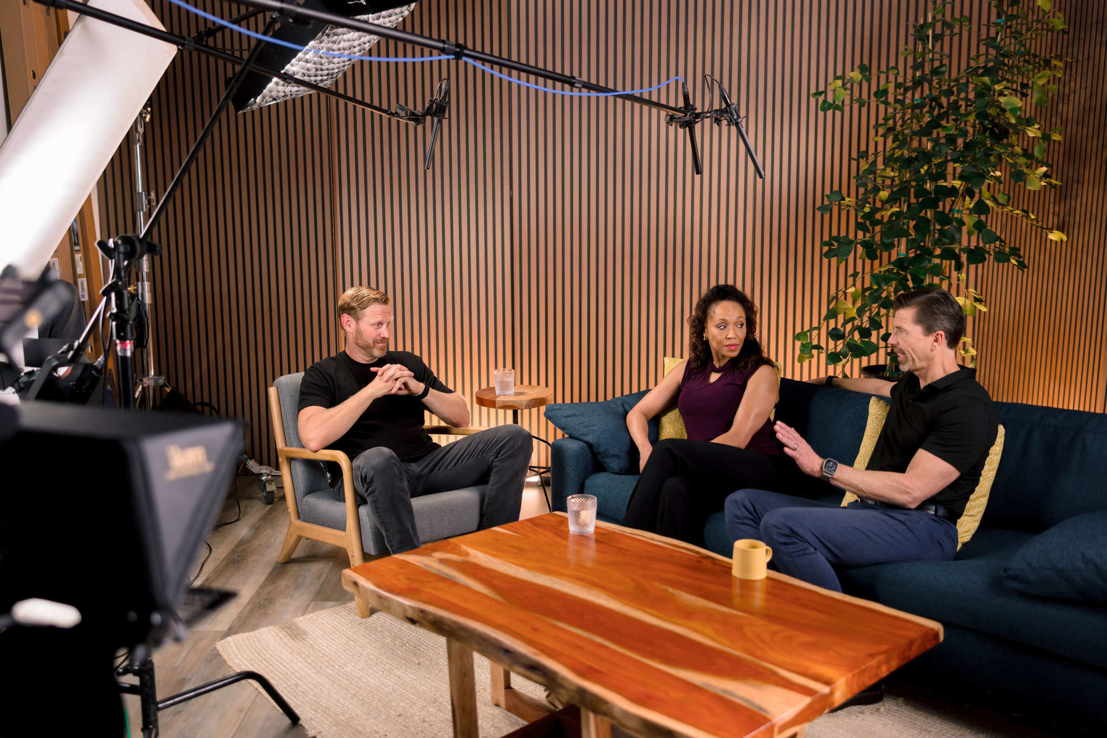 A well-lit studio interior features three seated individuals engaging in conversation, with a wooden table in front of them and microphones suspended overhead.