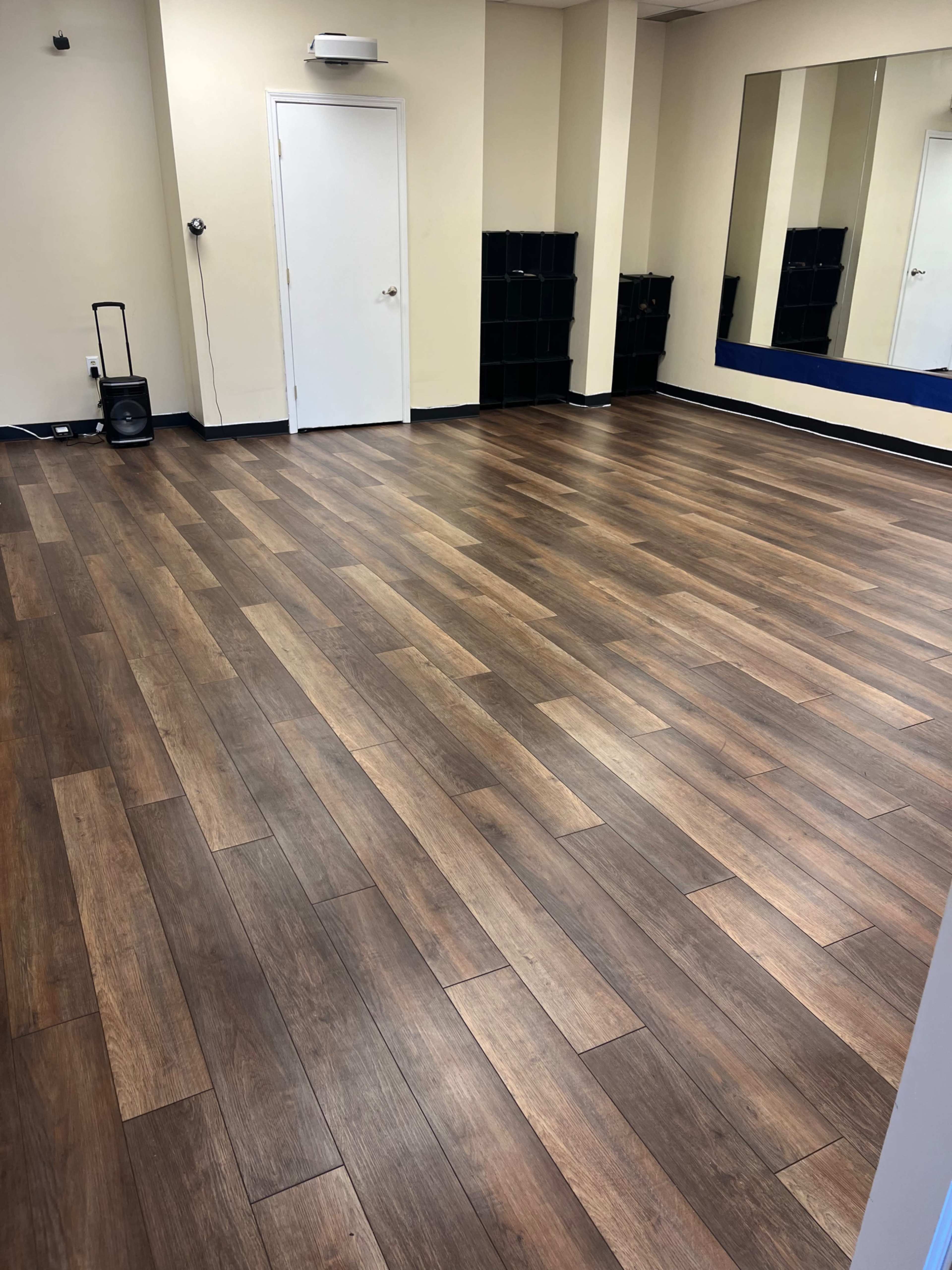 The image shows a spacious, empty room with wooden flooring, a mirror on the right wall, and a door in the back.