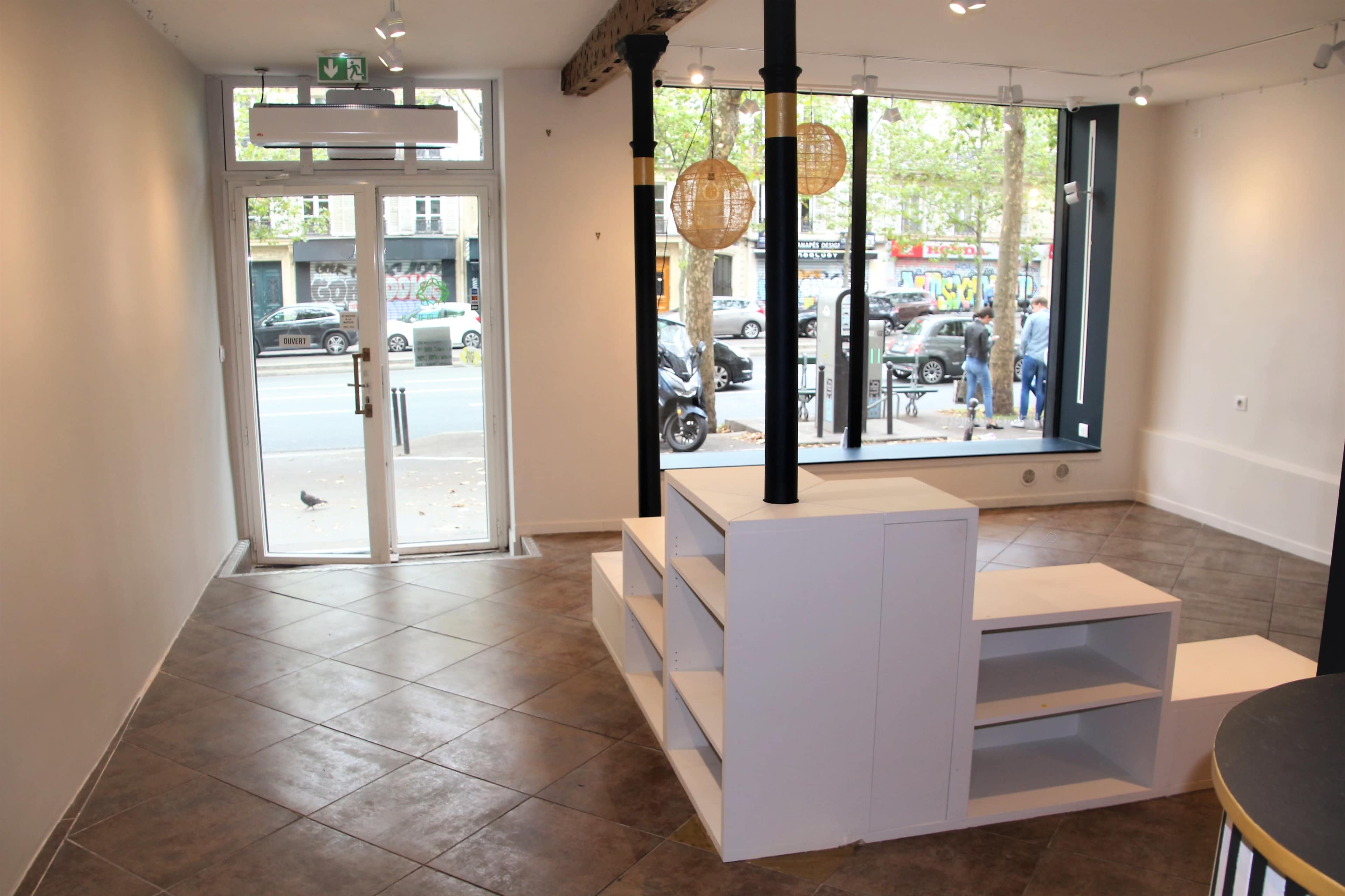 The image shows an empty room with a tiled floor, a large window displaying a street view, and a white shelving unit in the center.